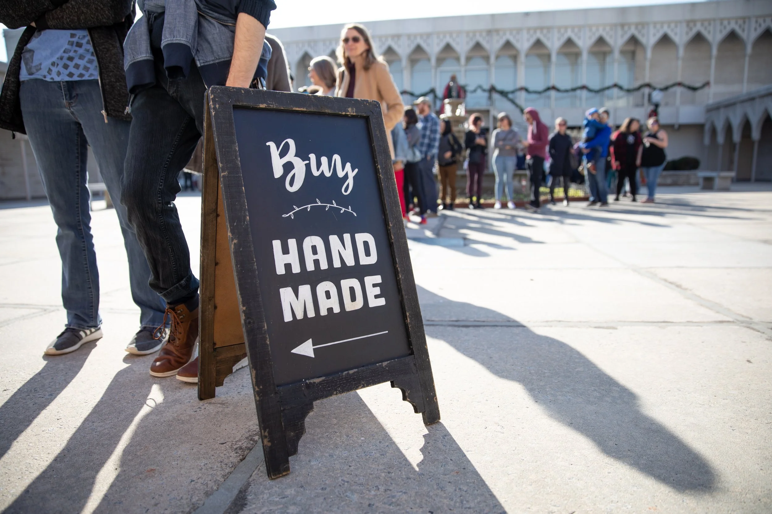 People standing in line outdoors near a sign that says 'Buy Hand Made' with an arrow pointing left, in front of a historic building.