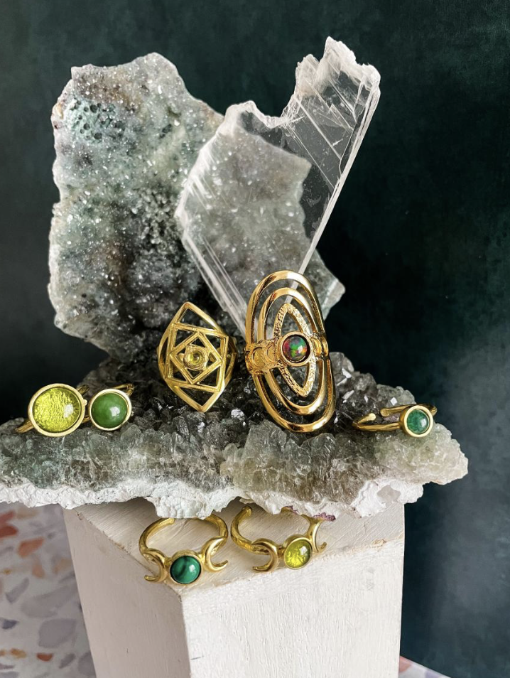Dream James Jewelery - Collection of gold jewelry with green and yellow gemstones displayed on a rock and white platform, with a piece of transparent mineral or glass in the background.