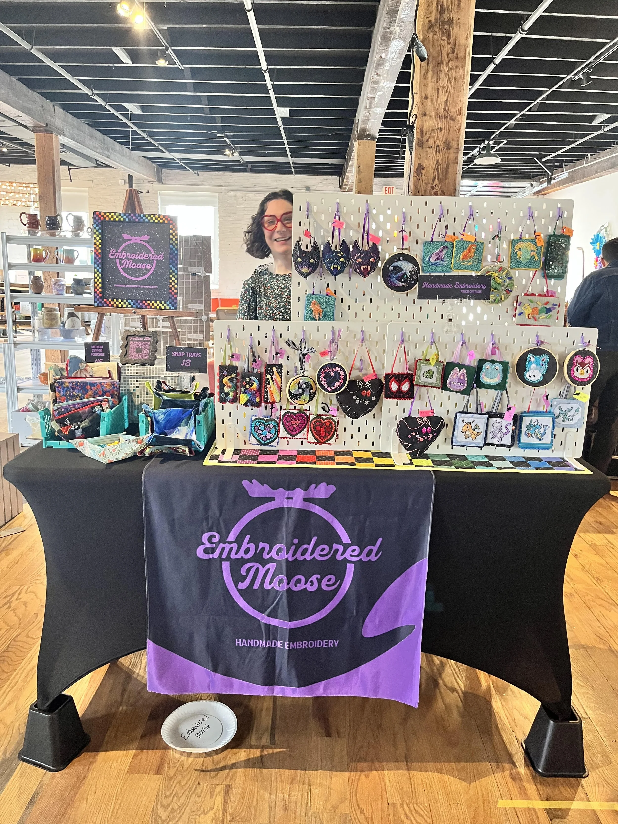 A table display at a craft fair with a purple and black cloth featuring the text 'Embroidered Moose' and a moose head logo. The table has various embroidered patches and items, including masks, bags, and keychains. A woman with dark curly hair, red glasses, and a floral top stands behind the display booth. The background shows a well-lit indoor space with exposed beams and other booths.