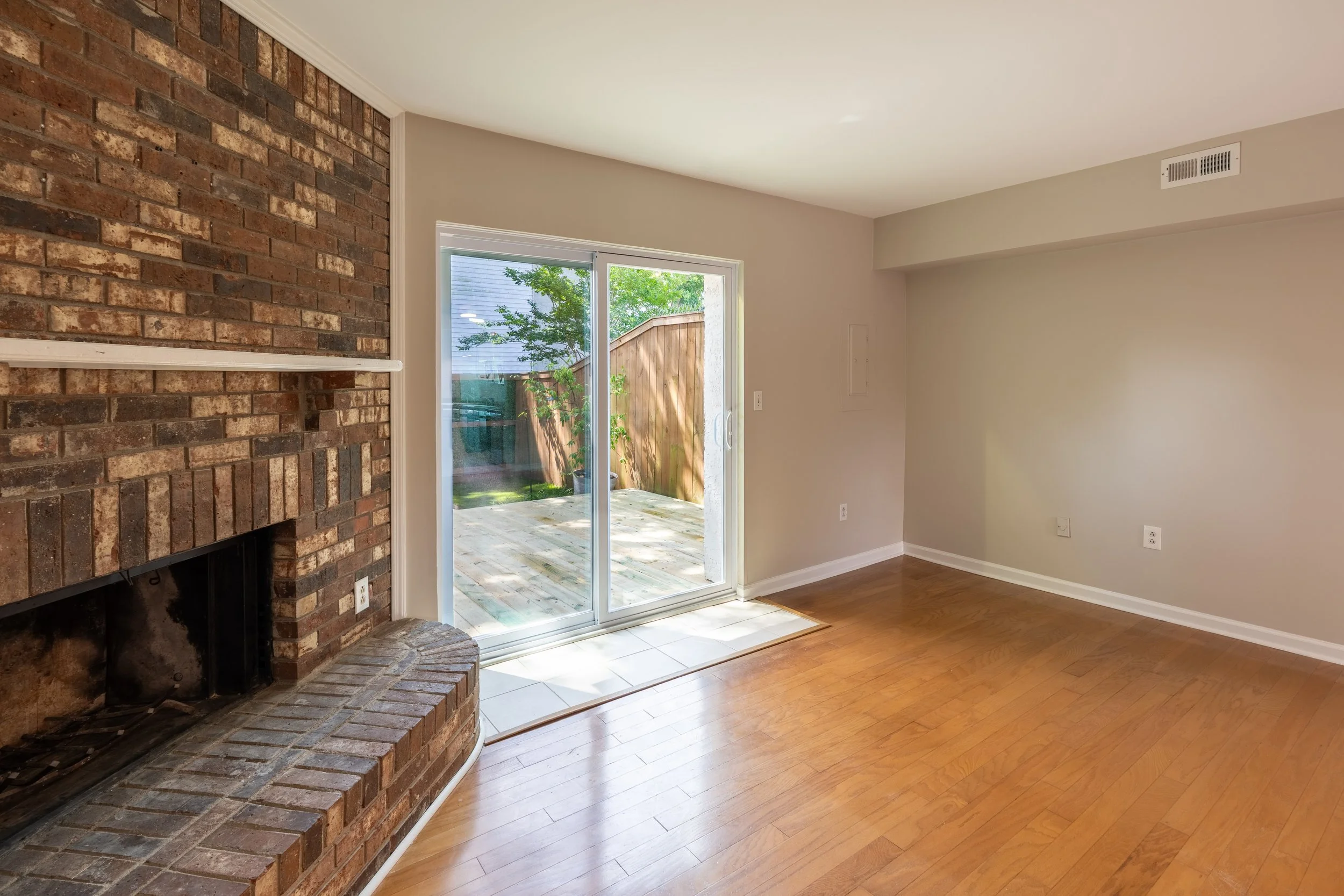 A living room with a brick fireplace, hardwood flooring, and a sliding glass door leading to a wooden deck outside. The room has beige walls and a vent near the ceiling.