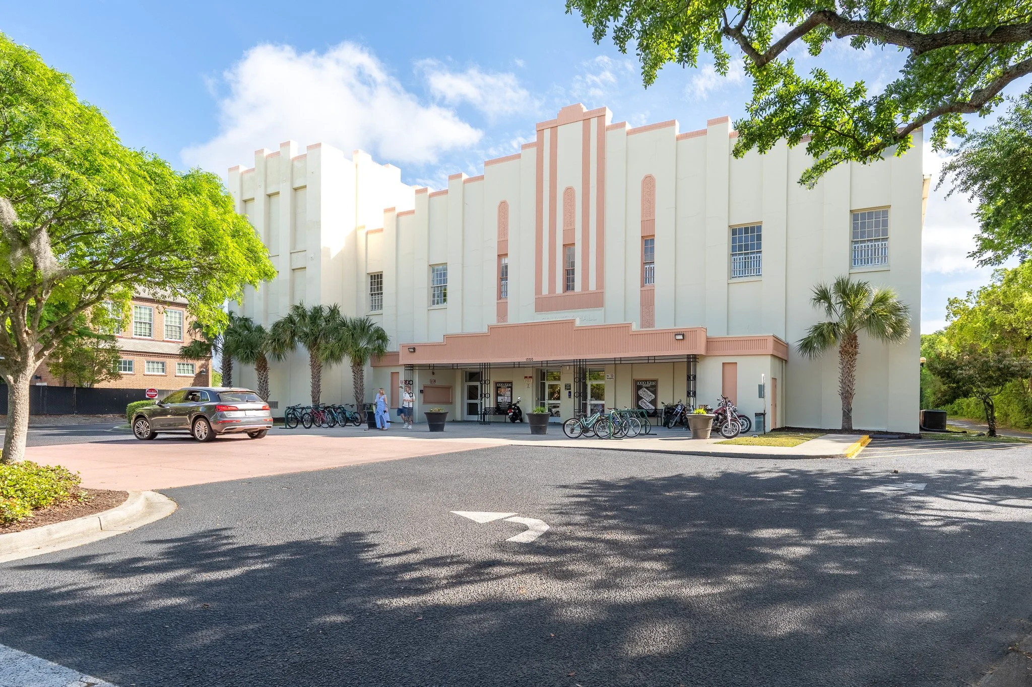 A white Art Deco style building with pink accents, surrounded by green trees, with parked bicycles and cars in front, under a partly cloudy sky.