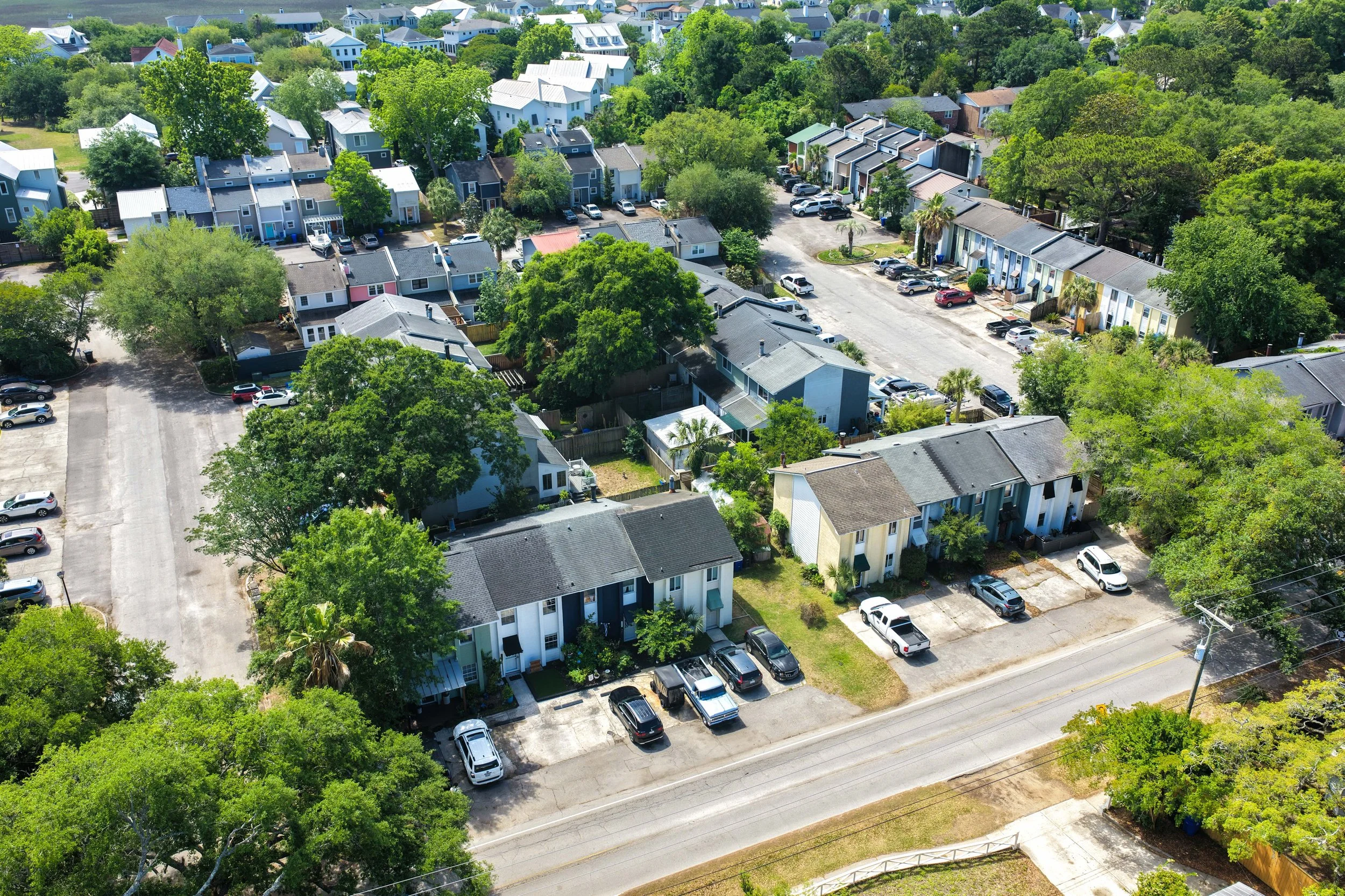 An aerial view of a residential neighborhood with rows of houses, green trees, and parked cars along the streets. There is a mix of single-family homes and townhouses, with some homes having small backyards and lush greenery.