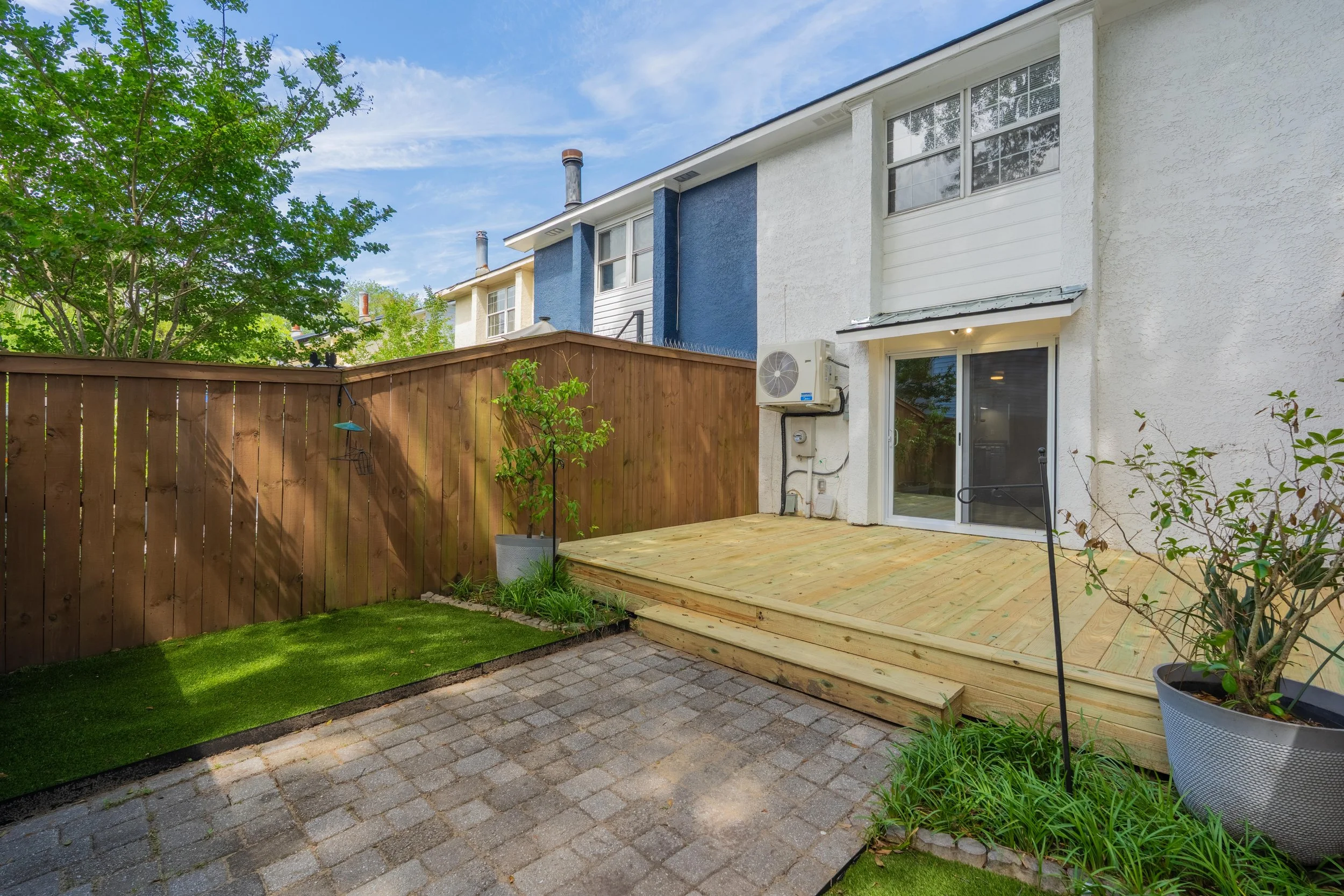 Backyard patio with a wooden deck, potted plants, a green lawn, a privacy fence, and the back of a house with large windows and sliding glass door.