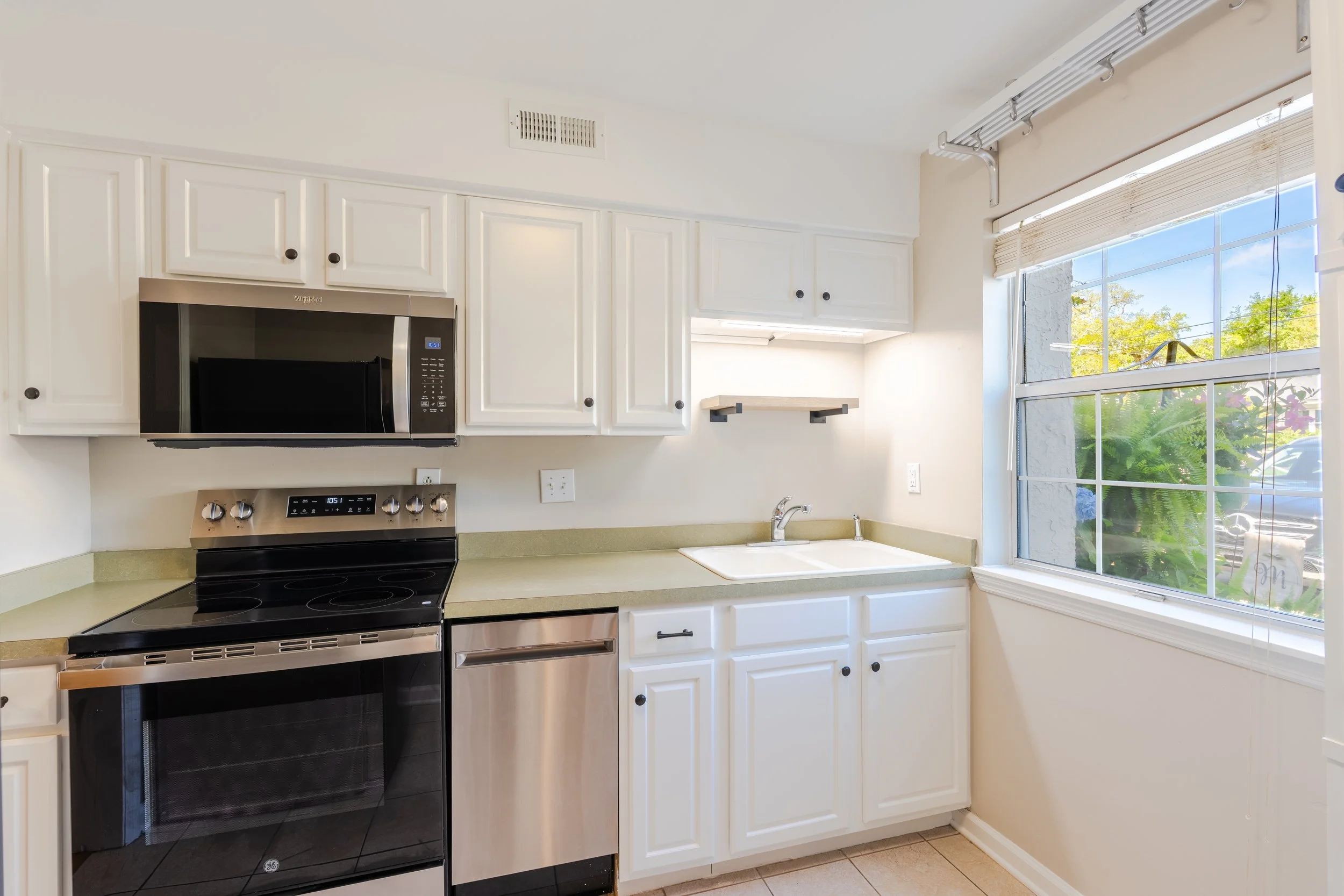 A kitchen with white cabinets, a black microwave oven, stovetop, and a yellowish-green countertop. There is a window with blinds and a view of trees outside.