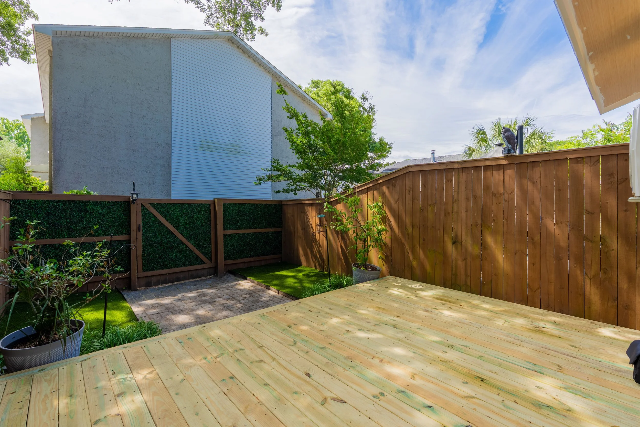 A partially constructed wooden deck in a backyard with a wooden fence, potted plants, a small grassy area, and a large tree. In the background, there is a neighboring house with white siding and a blue sky with some clouds.