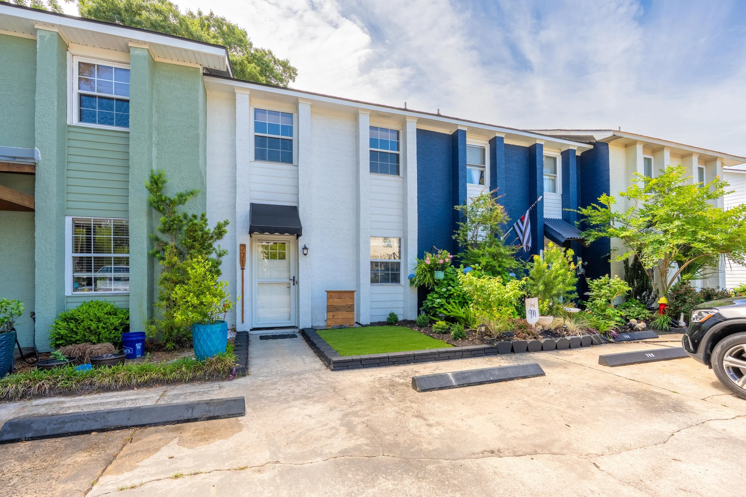 Colorful multi-story residential building with painted exterior walls in green, white, and blue, separated by white vertical columns. The building has several windows, some with blinds and some open. There is a small front yard with green plants, tre