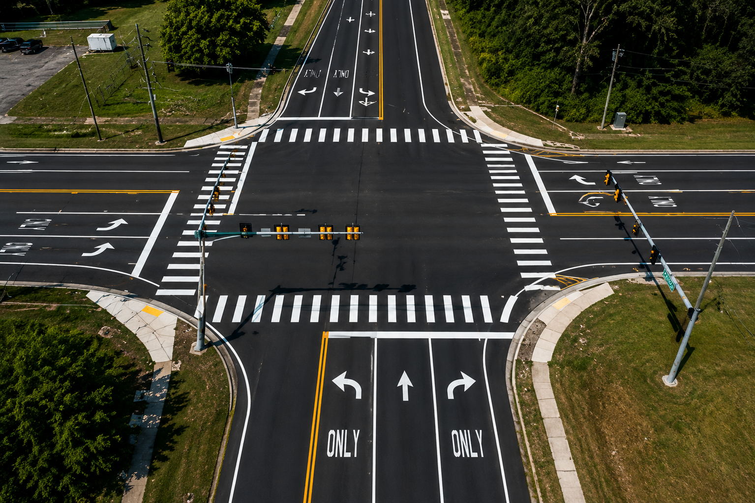 A newly paved intersection with multiple lanes, traffic signals, and painted arrows indicating directions for vehicles, surrounded by grassy areas and trees.