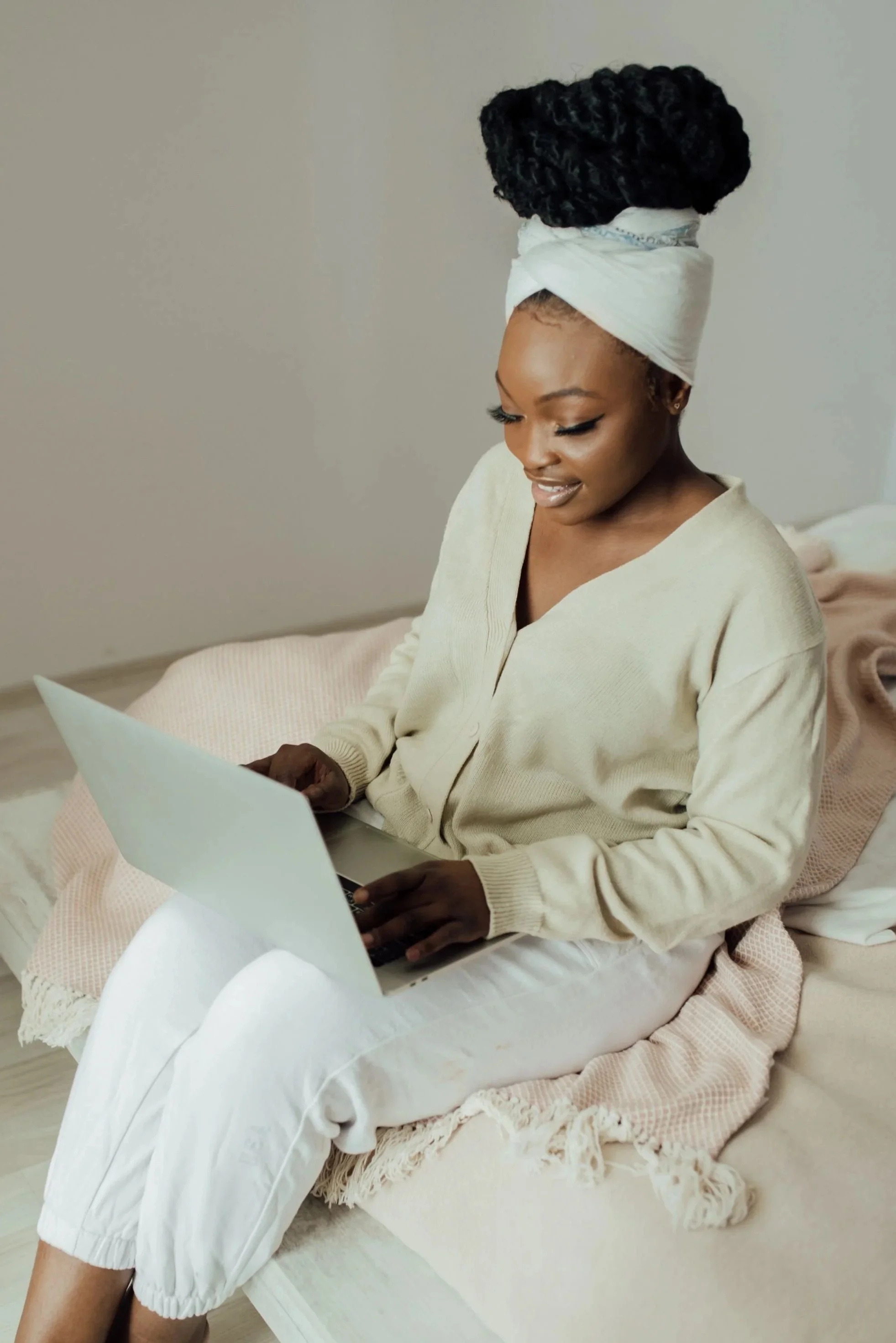 Woman with styled curly hair wrapped in a white cloth tied in a turban, sitting on a bed, using a laptop
