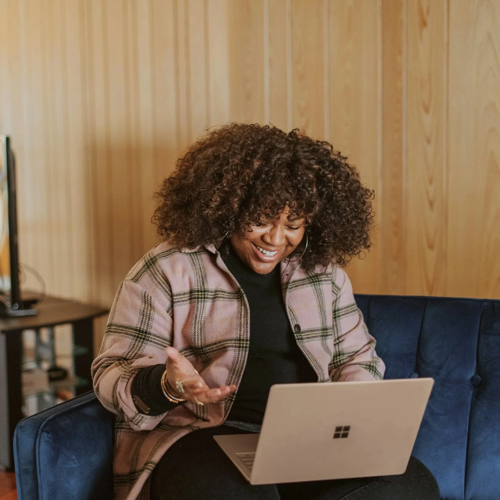 Woman with curly hair smiling and talking to someone while sitting at a table with a laptop.