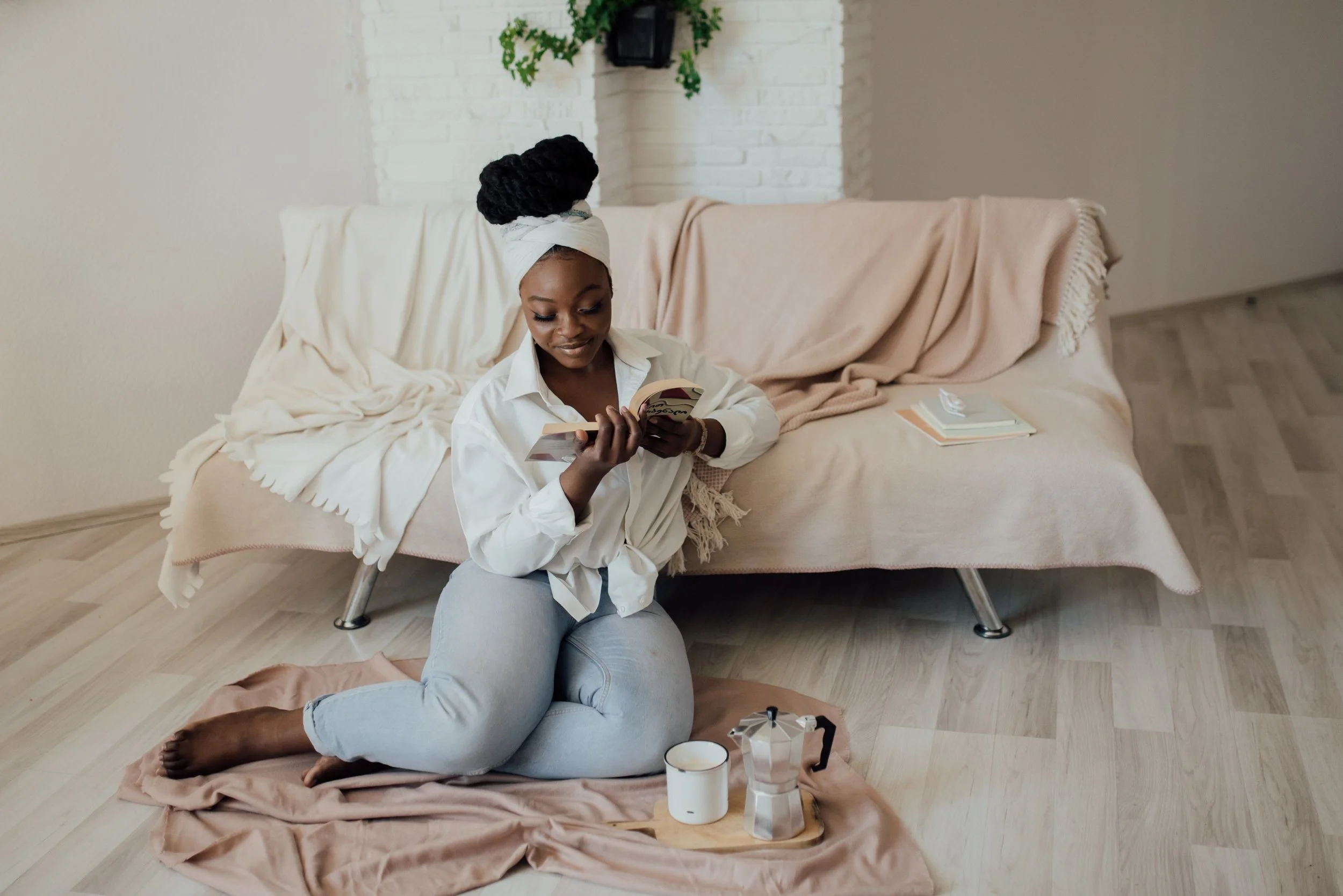A woman sitting on the floor in front of a couch, reading a book. She is wearing a white shirt and gray pants, with her hair wrapped in a white headscarf. There is a coffee mug and a French press on a blanket on the floor nearby. The couch has a beige throw blanket draped over it, and there are some books on the floor next to it. The background features a white brick wall with a small plant hanging from the ceiling.