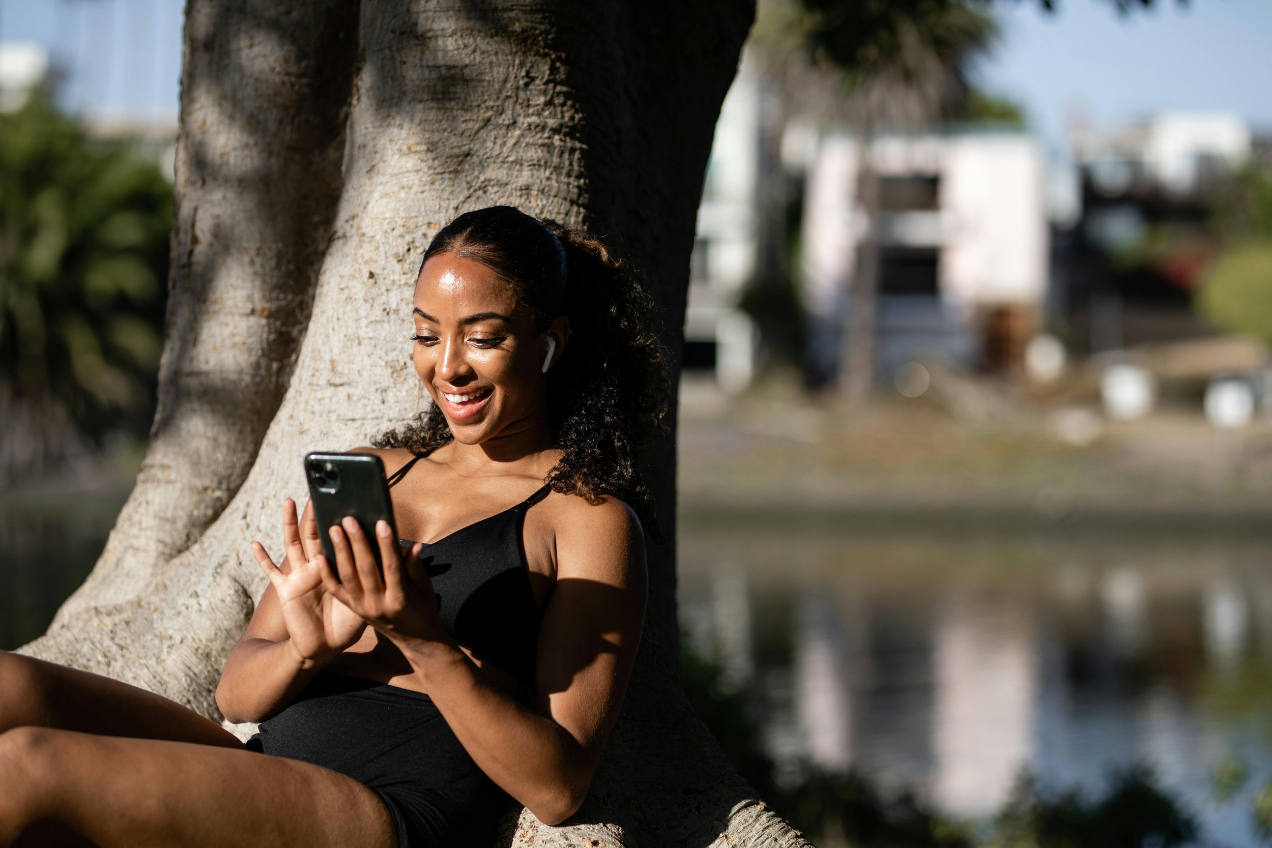 A woman with curly hair sitting outside by a tree, smiling and looking at her phone.