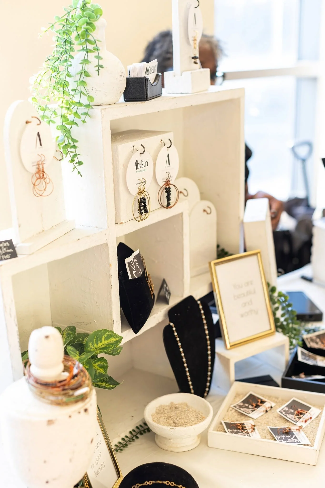 Display of jewelry including earrings, necklaces, and bracelets on a white shelf with decorative plants and framed signs, inside a well-lit shop or market.