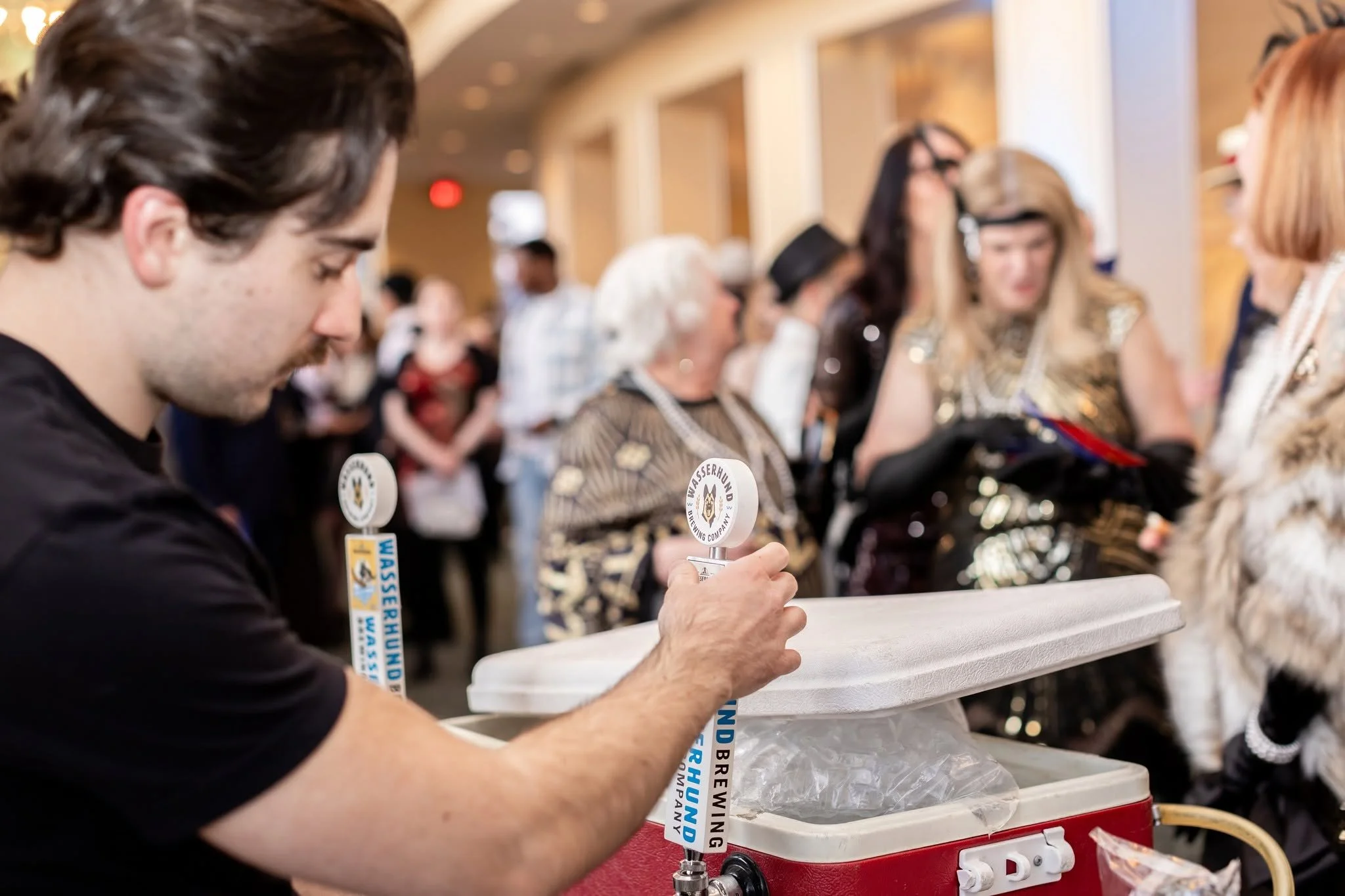 A man is filling a cup with beer at an event, with people in the background. The scene is lively with several attendees, and the man is focused on pouring the beer.