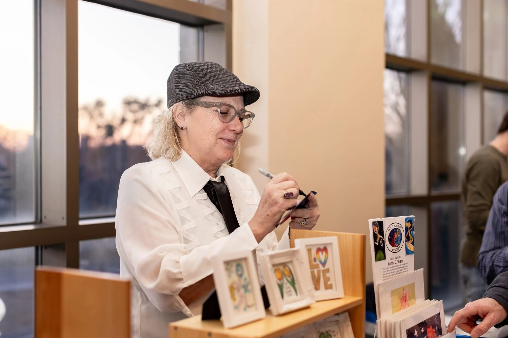 A woman with blonde hair, wearing glasses, a gray hat, a white jacket, and a black tie, is standing at a table with books and cards, writing on a notepad.