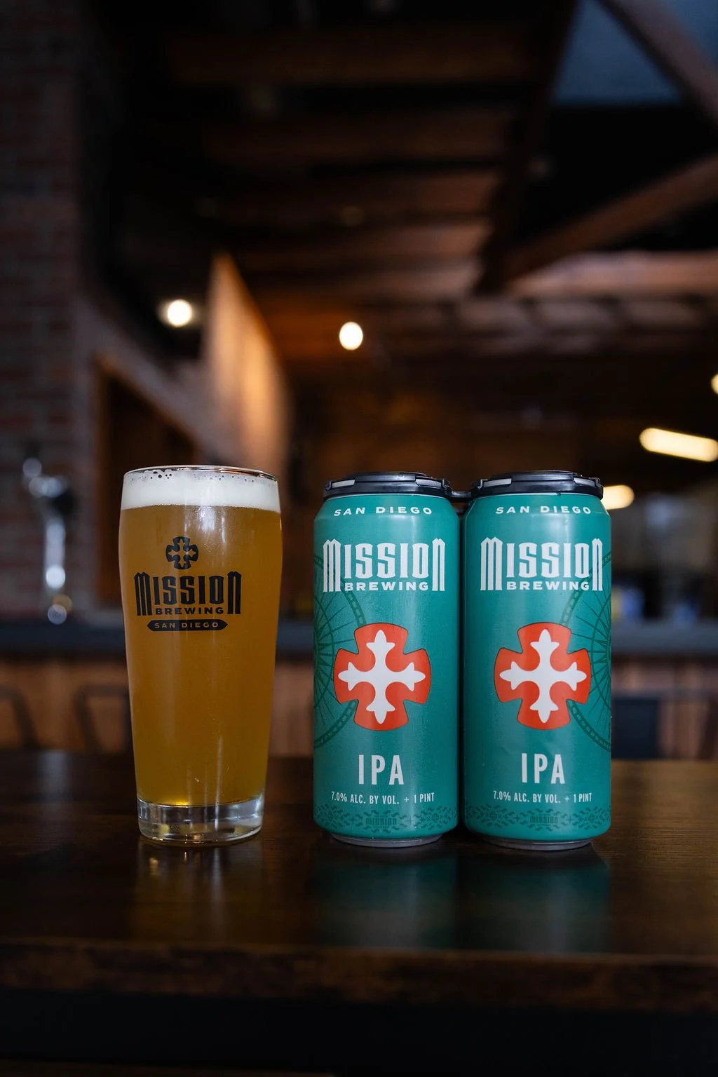 A pint glass filled with pale golden IPA beer with a foam head, labeled 'Mission Brewing San Diego', next to two teal cans of Mission Brewing IPA on a wooden table indoors.