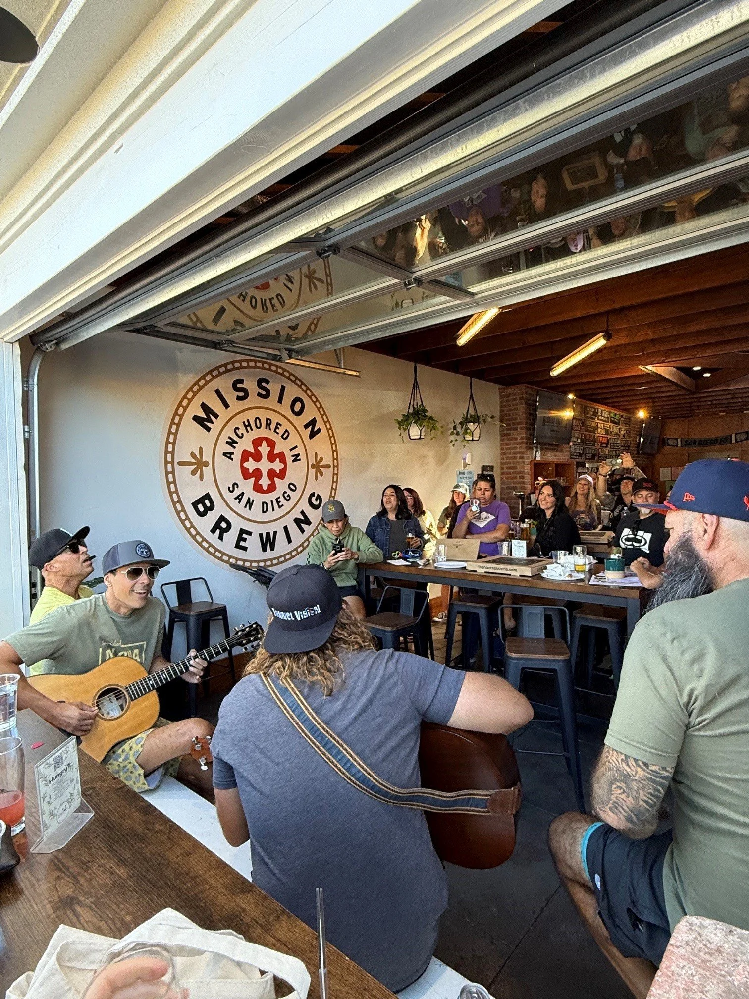 People in lively Mission Brewing Kensington, one person playing guitar, others singing and socializing, with Mission Brewing logo on the wall and a mix of seated and standing guests.