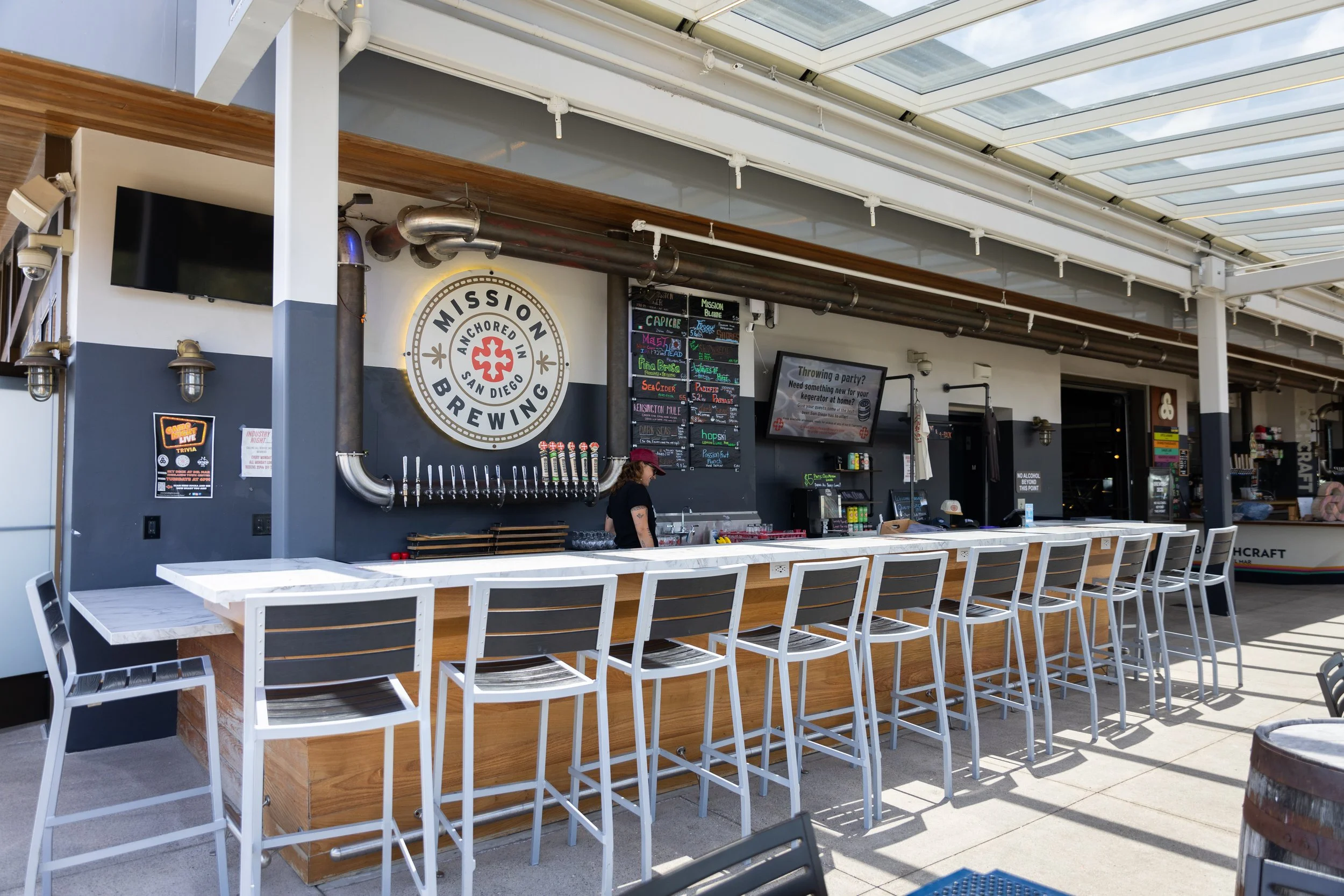 A bar area in a brewery with a white marble countertop, high chairs, and a wall sign that reads 'Mission Brewing Anchored in San Diego.' There are several taps on the wall, digital menu screens, and a person wearing a red cap behind the bar. The space has a modern industrial design with exposed pipes and a skylight ceiling.