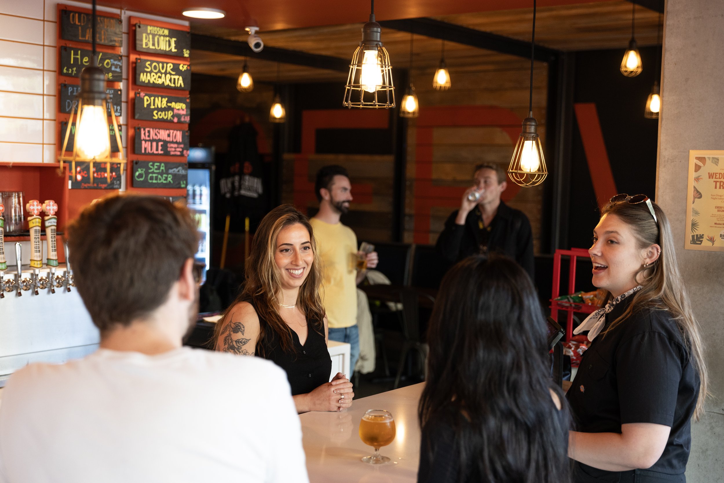 People gathered around a bar counter in a Mission Brewing UCSD, some people are smiling and talking, while a woman with a tattoo on her arm appears to be engaged in conversation.