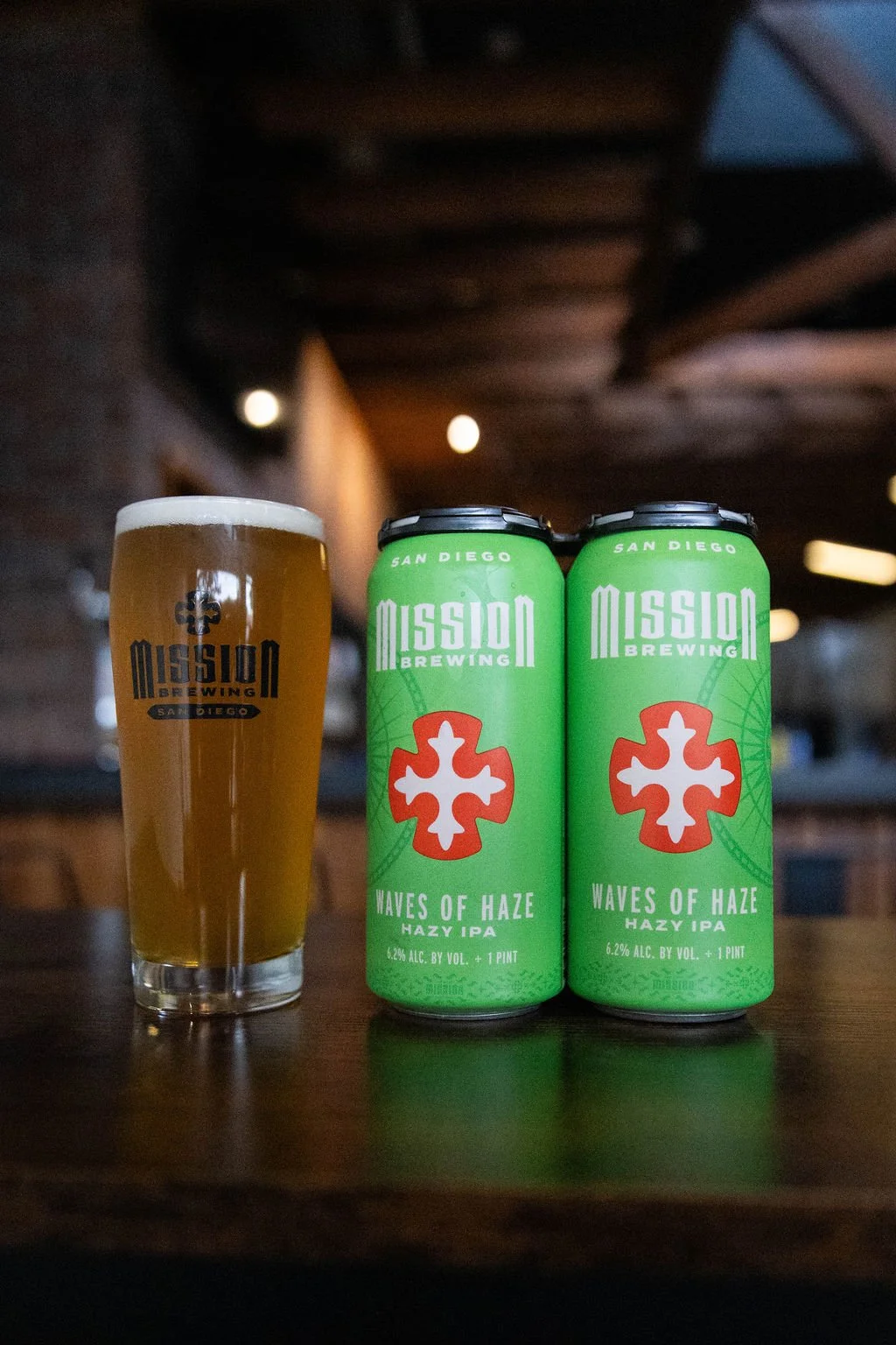 A pint glass filled with beer and two cans of Waves of Haze Hazy IPA from Mission Brewing on a wooden table indoors.