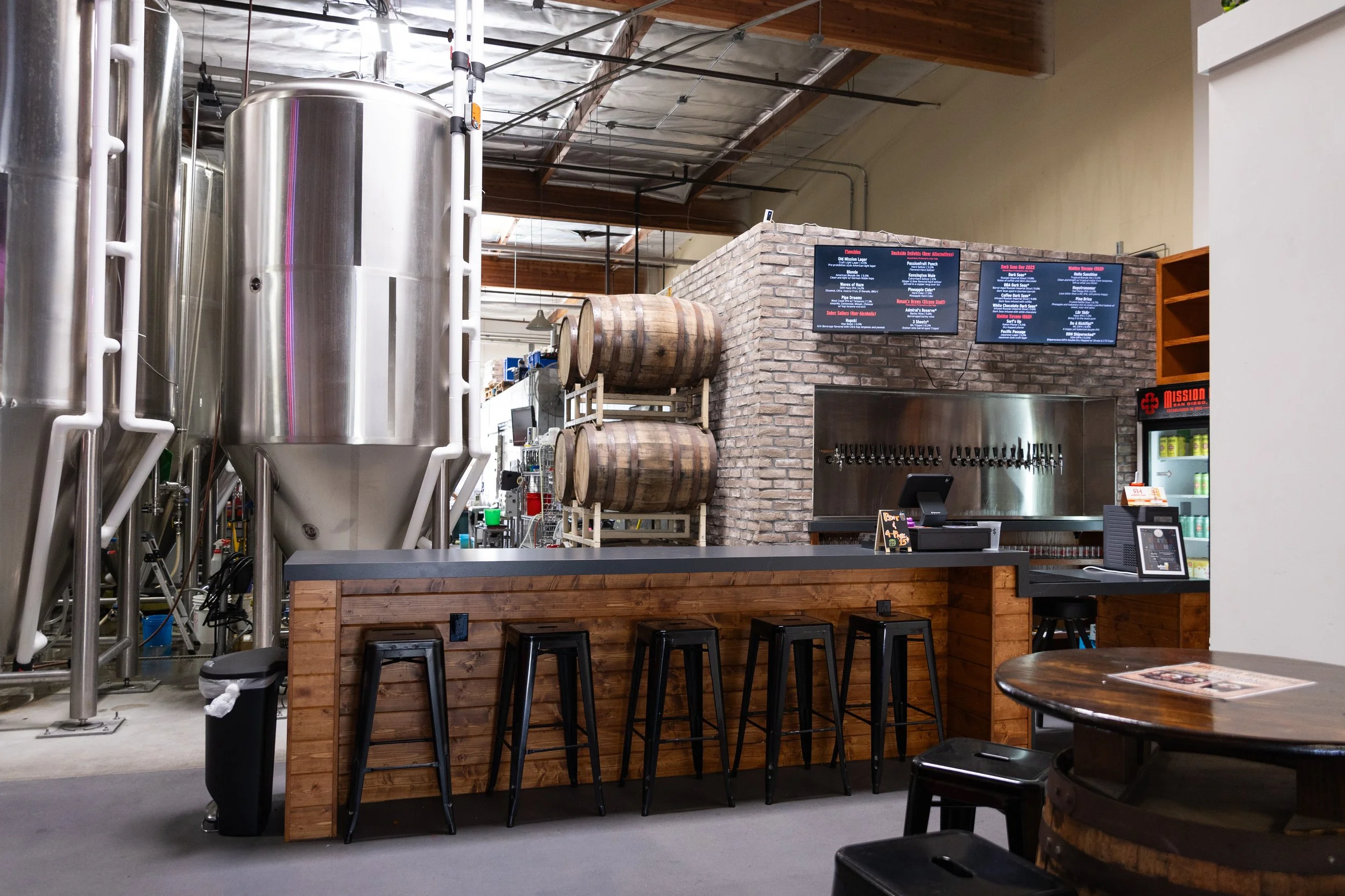Inside a brewery or taproom featuring large stainless steel brewing tanks, wooden barrels, brick walls with digital menu screens, a counter with bar stools, and Tables with chairs.