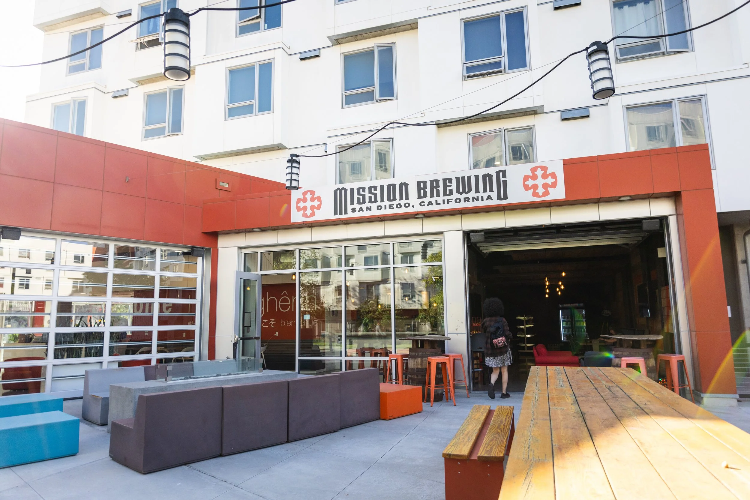 Outdoor patio of Mission Brewing in San Diego, California, with seating area, tables, and people near the entrance. Modern building with multiple windows in the background.