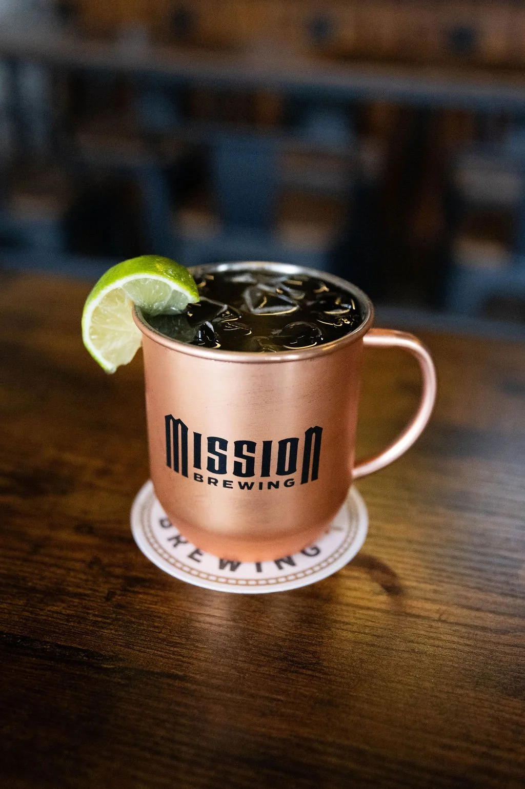 A copper-colored mug with 'Mission Brewing' logo, filled with a dark beverage and ice, garnished with a lime wedge, sitting on a wooden table.
