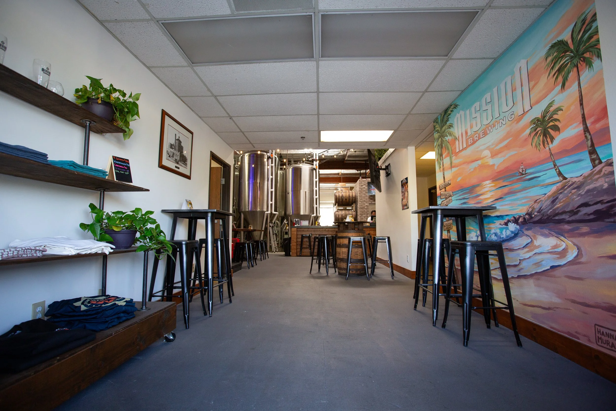 Interior of a brewery or taproom with high tables and barstools, a large colorful wall mural of a beach scene with palm trees, and brewing equipment visible in the background.