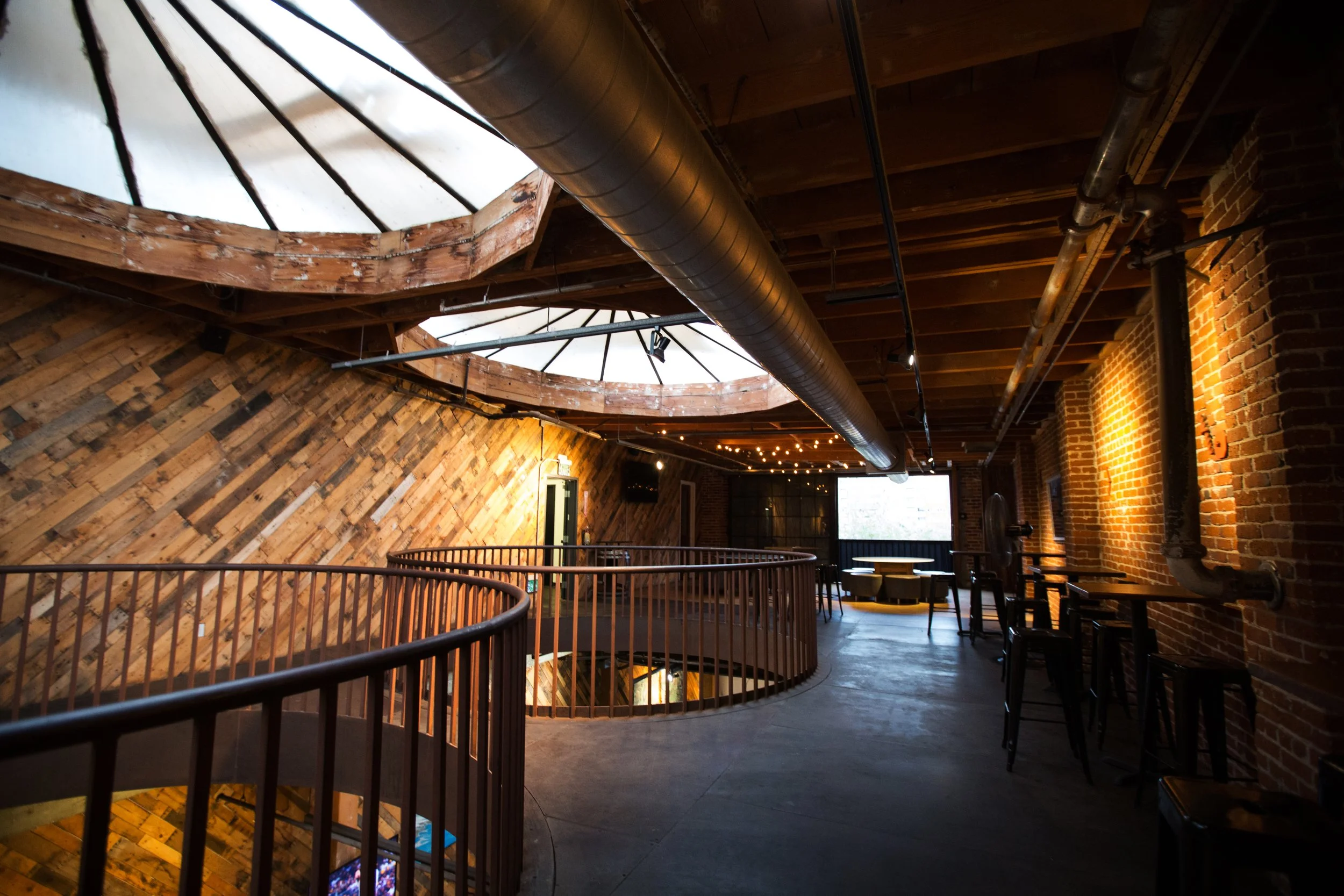 Interior of a loft-style space with brick and wooden walls, visible ductwork, a balcony with a metal railing overlooking a lower level, windows letting in natural light, and modern furniture including chairs and tables.