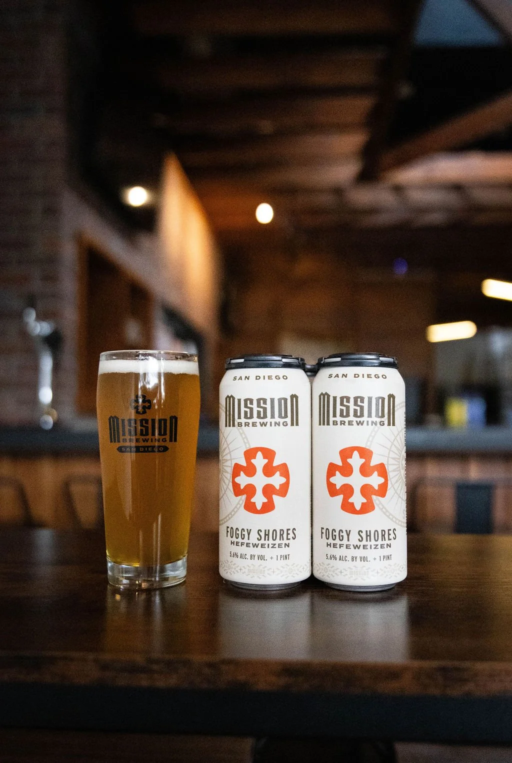 Two cans of Mission Brewing Foggy Shores Hefeweizen beer and a glass filled with beer on a wooden table indoors.