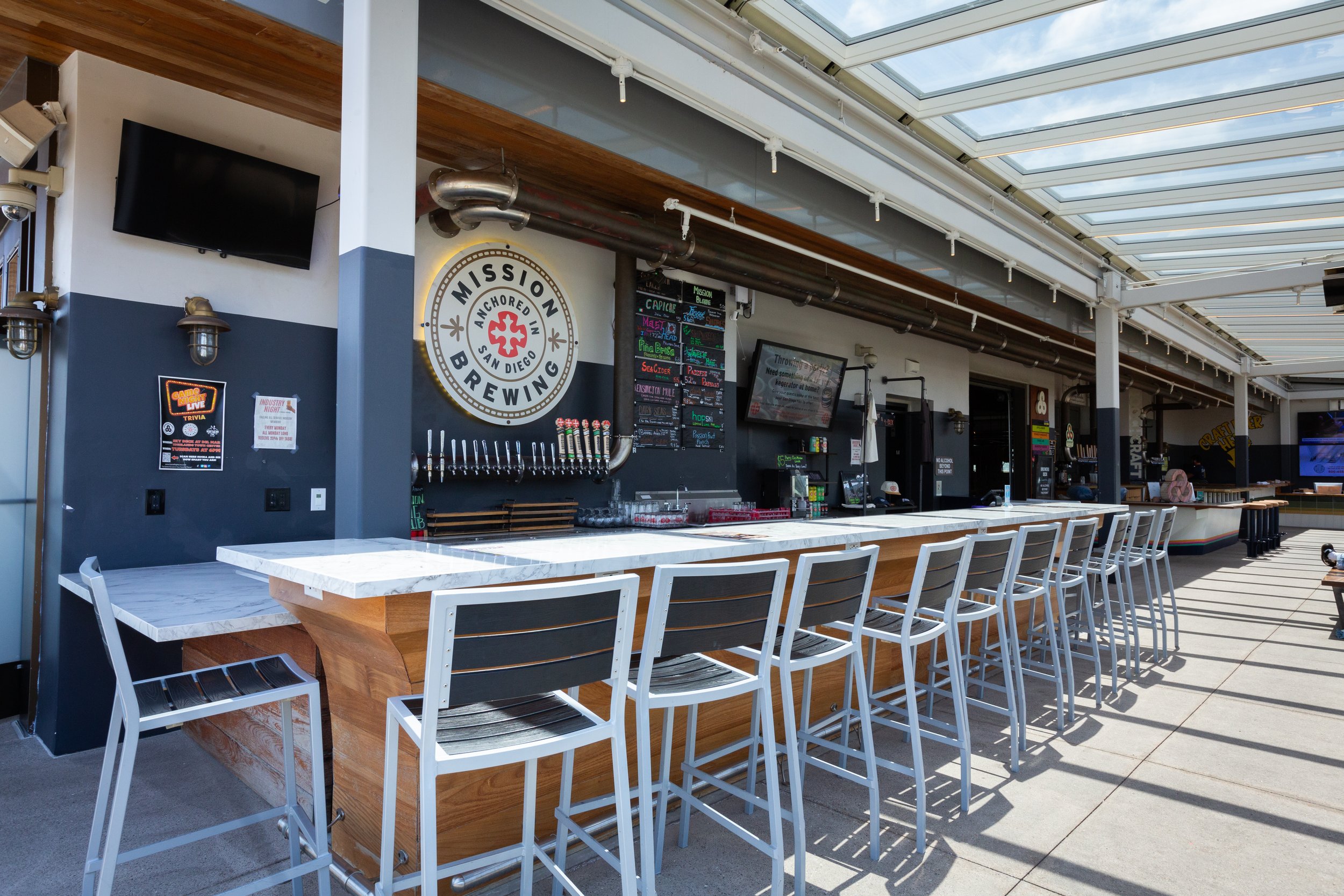 Bar area at Mission Anchor Brewing in San Diego with white chairs, a bar with taps, and chalkboard menus, under a glass roof.