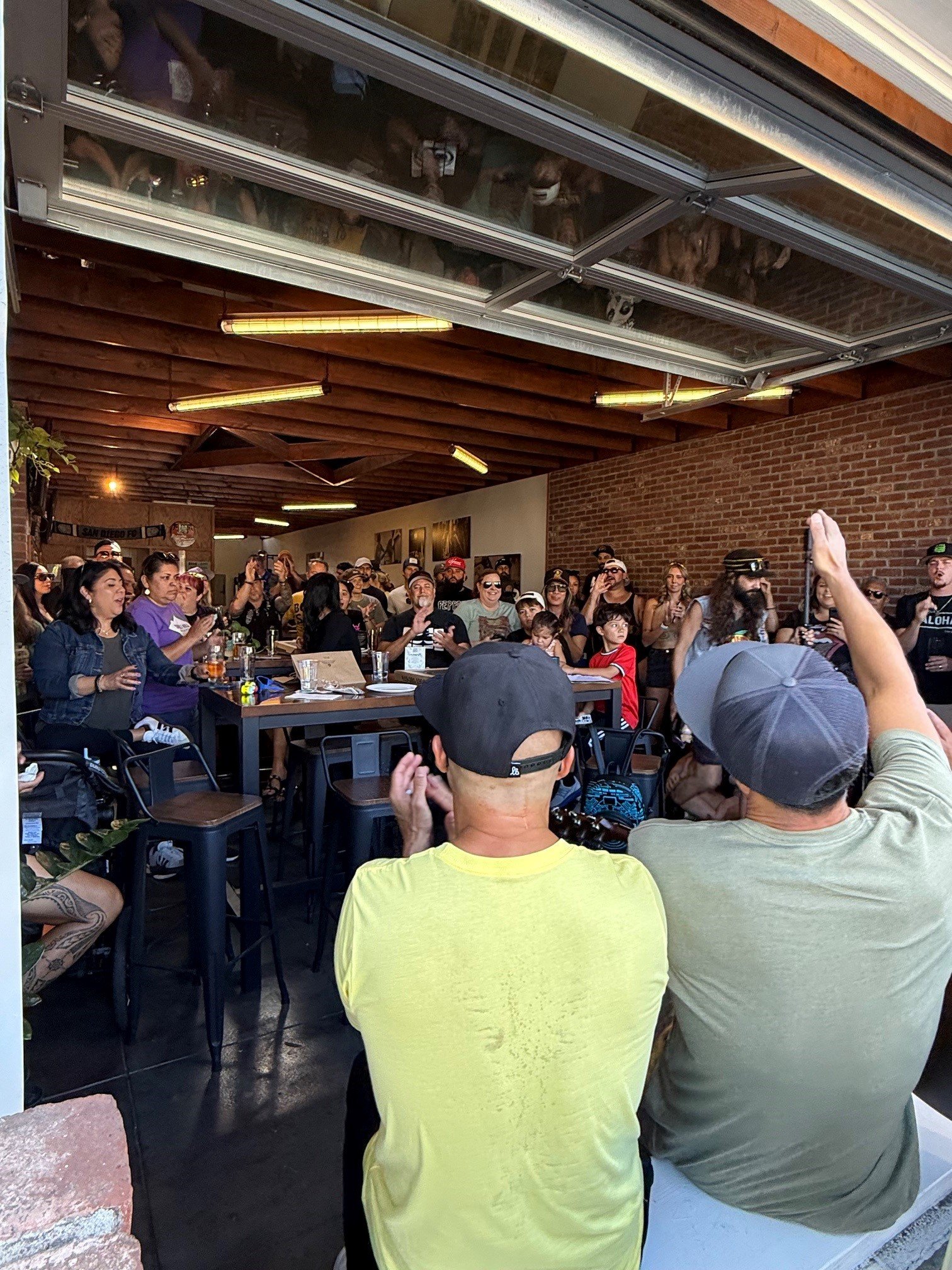 A crowded indoor gathering at Mission Brewing Kensington with people of various ages, some wearing sunglasses and hats, socializing and sitting at tables in a restaurant or event space with brick walls and wooden ceiling beams.