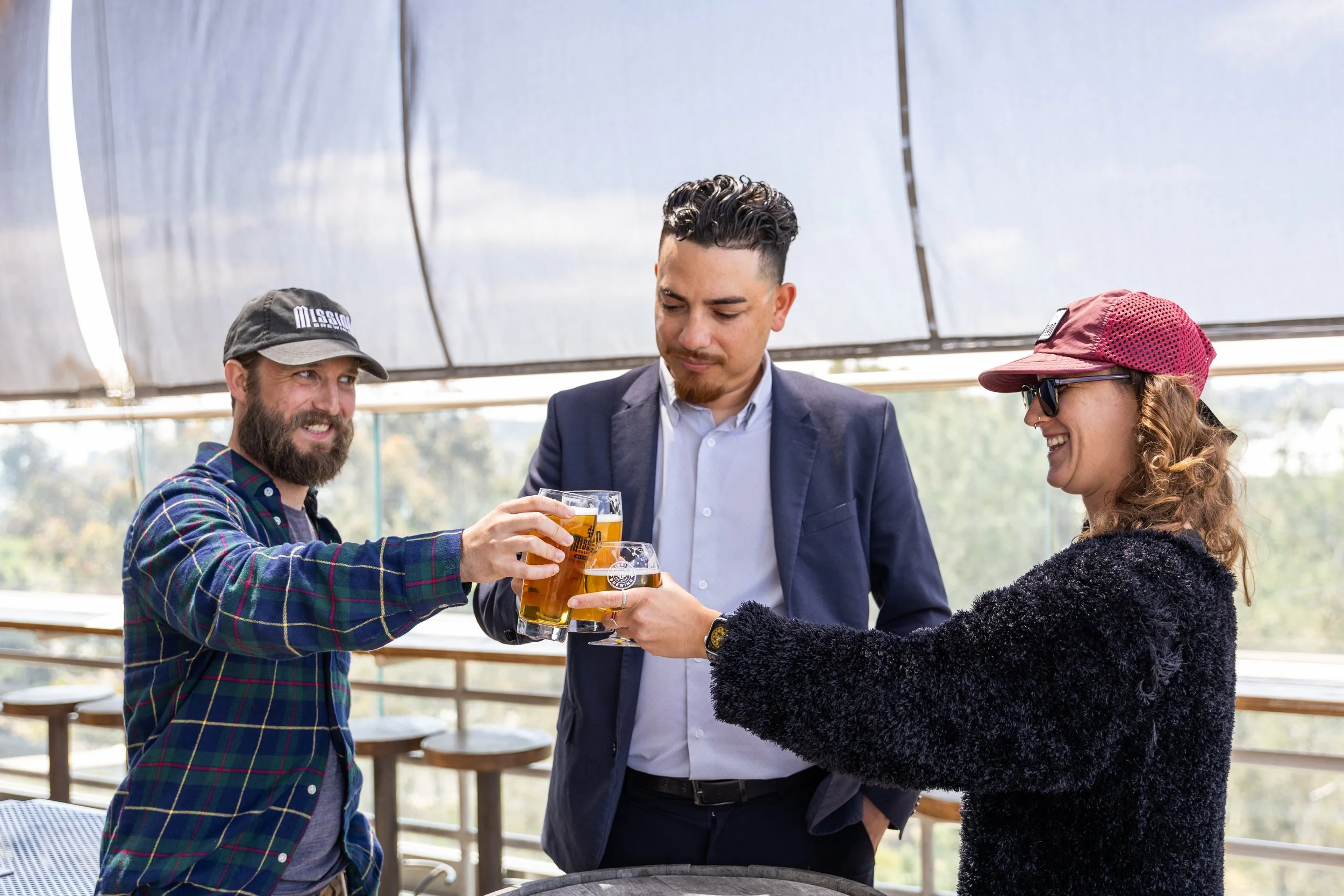 Three people at an outdoor Mission Brewing patio clinking beer glasses, two men and one woman, with smiles, under a shade structure, with trees and sky in the background.