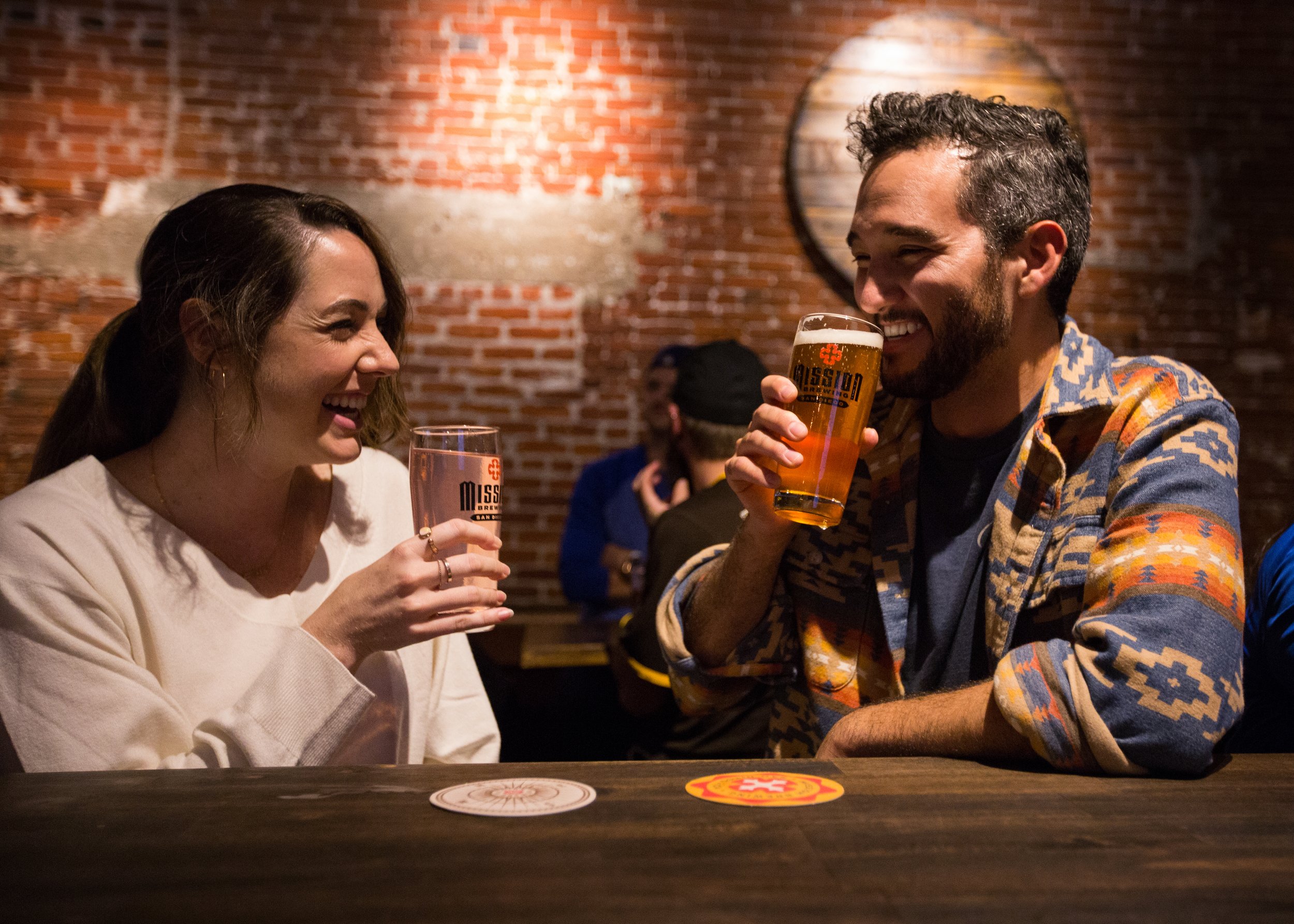 Two people, a woman and a man, are laughing and enjoying drinks at a bar. The woman has long dark hair and is wearing a light-colored top, while the man has curly dark hair and a beard, wearing a colorful patterned shirt. They are holding glasses of 