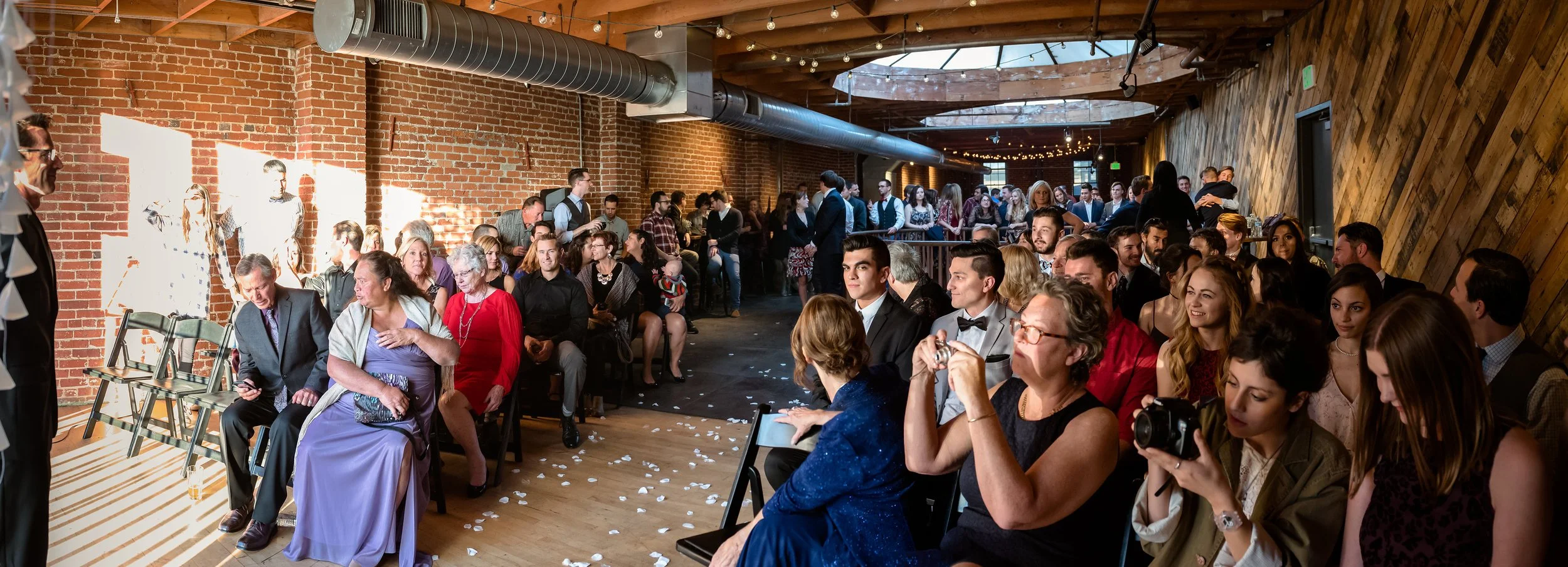 A crowded indoor event with many seated and standing people, some dressed formally. The room has exposed red brick walls, wooden accents, and string lights hanging from the ceiling. The sunlight streams through a window, illuminating part of the room.