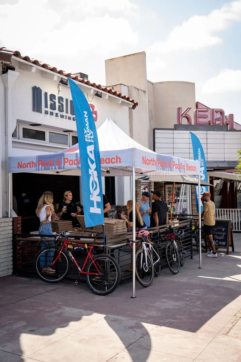 People waiting in line at a food pickup stand under a white canopy with blue flags that read "HOVAN" and "North Park & Pacific Beach." Bicycles are parked in front of the stand. In the background, there are buildings and a marquee with pink letters.