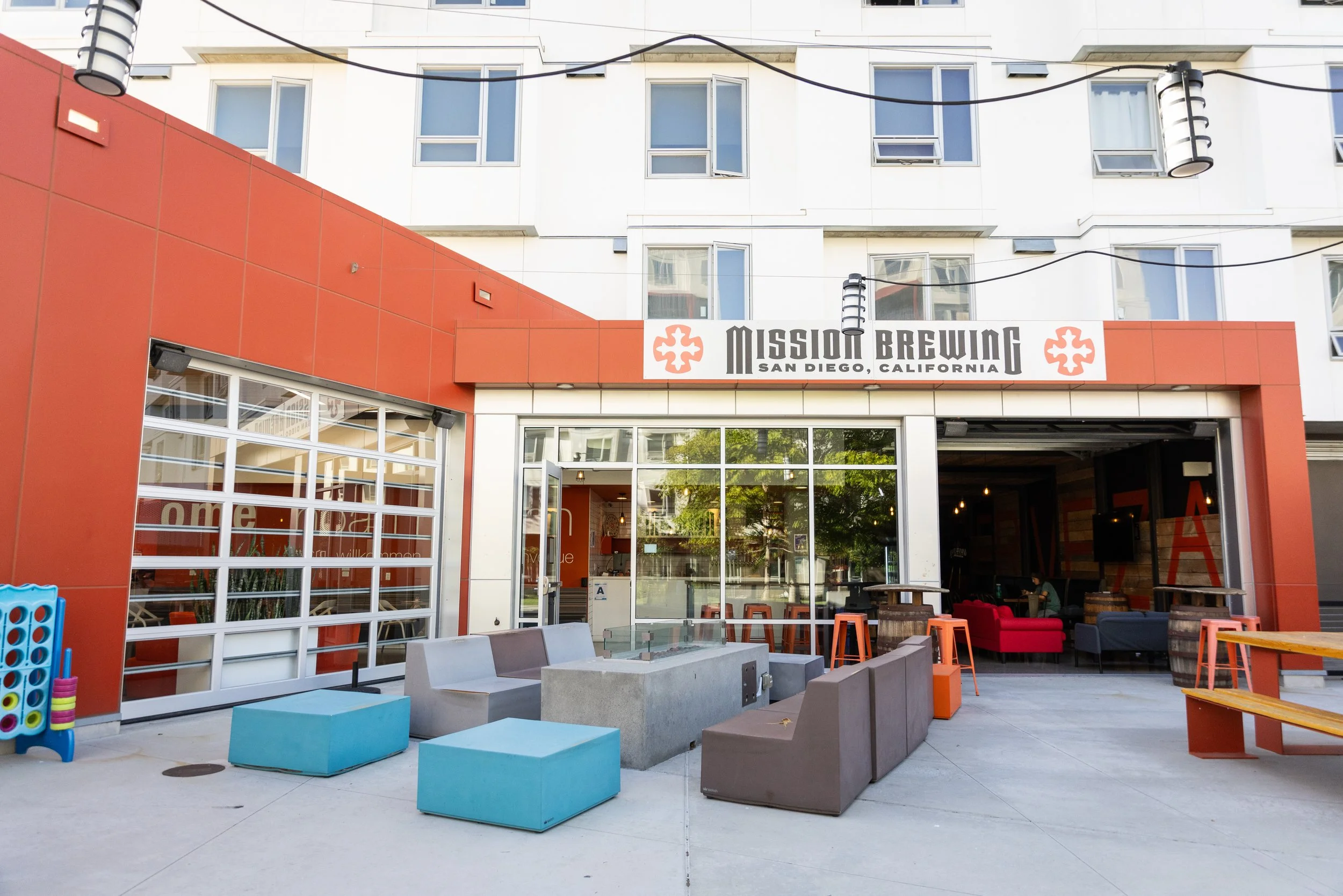 Outdoor seating area of Mission Brewing in San Diego, California, featuring colorful chairs, couches, tables, and playful design elements in front of a modern building with large windows and a red accent wall.