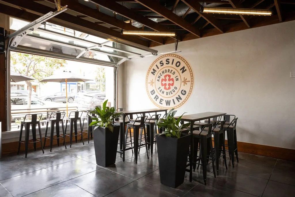 Interior of Mission Brewing Kensington with high tables, chairs, and a large window showing outdoor chairs and umbrellas.