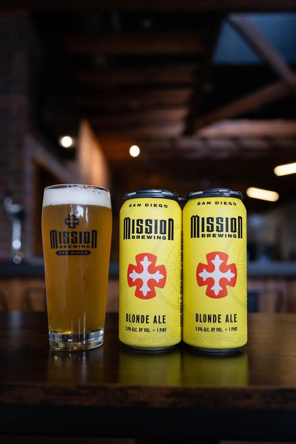 A glass of Blonde ale beer with Mission Brewing logo next to two cans of Mission Brewing Blonde Ale on a wooden table indoors.