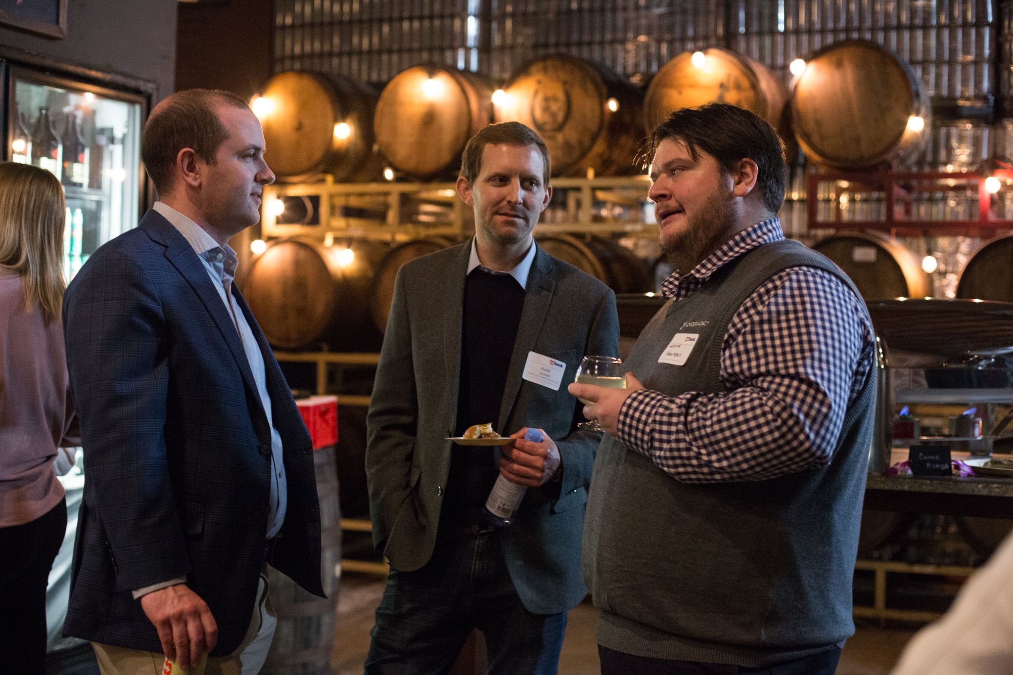 Three men having a conversation at a social gathering in a Mission Brewing East Village, with large barrels in the background and warm lighting.