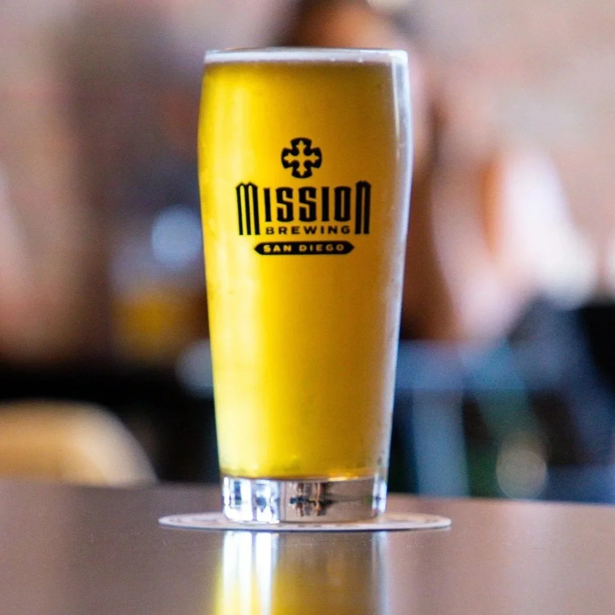 A tall glass of beer with the logo of Mission Brewing in San Diego, featuring black text and a cross-like symbol on a golden beer background.