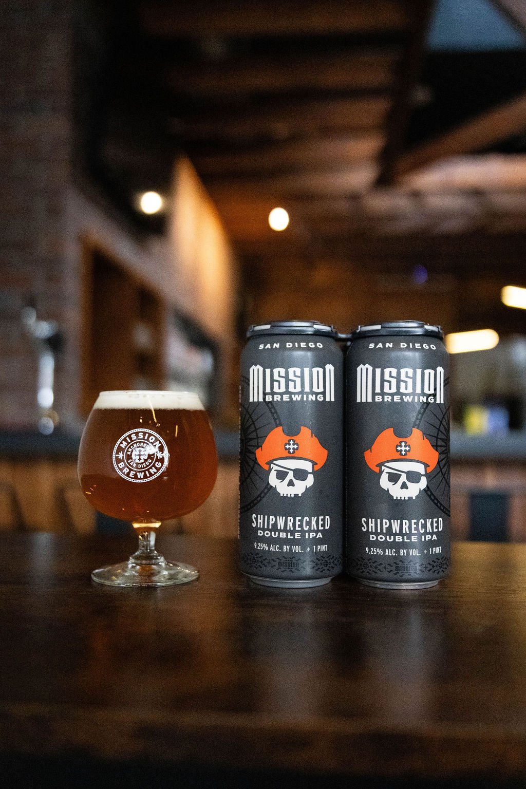 Two cans of Mission Brewing Shipwrecked Double IPA beer and a snifter glass of beer on a wooden table indoors.