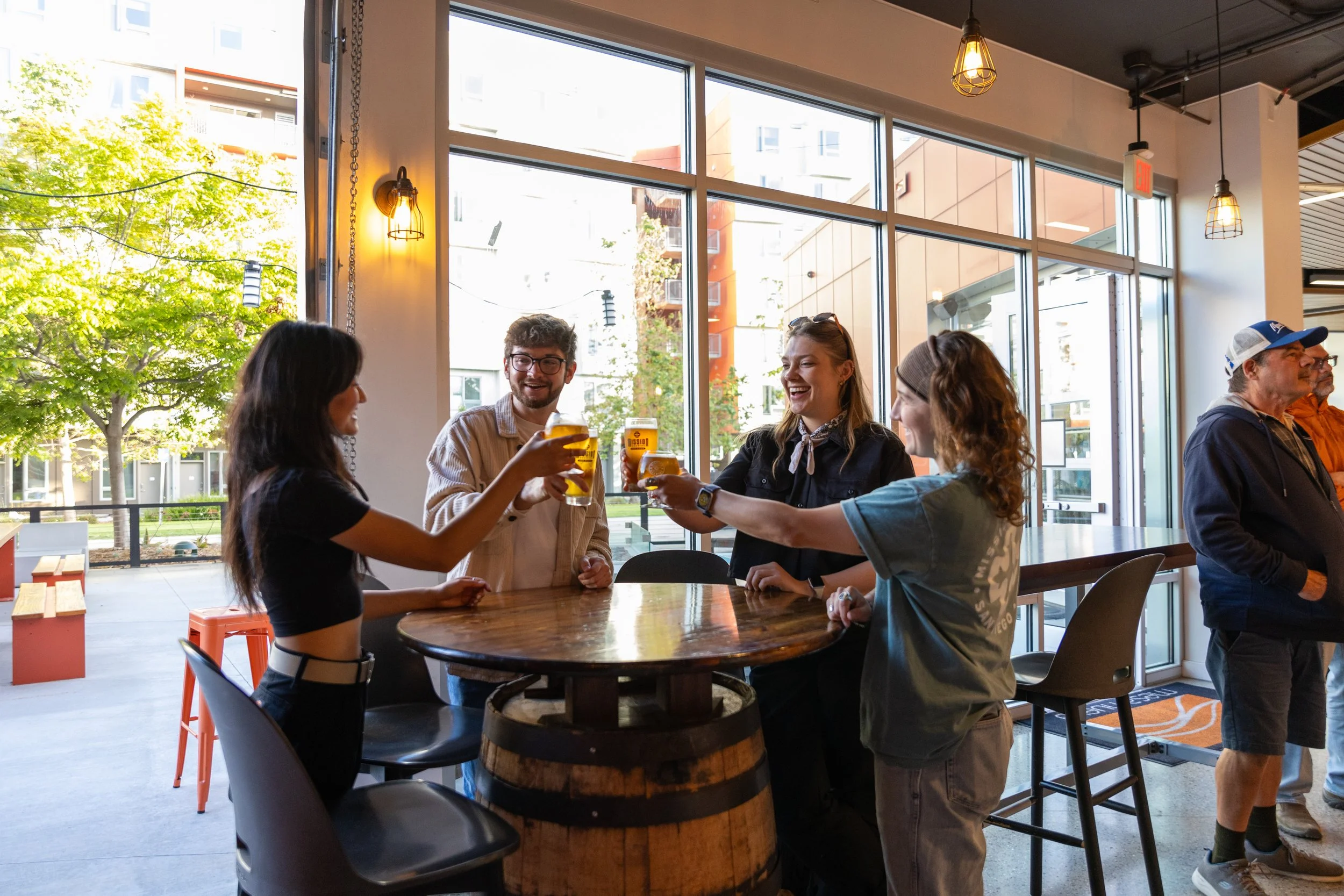 Group of five people standing around a wooden table inside a Mission Brewing UCSD, holding drinks and talking, with large windows showing trees and buildings outside, and warm indoor lighting.