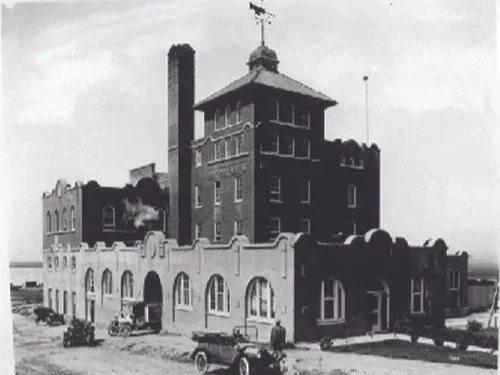 Black and white photo of a large, multi-story brick building with a tower, arched windows, and a chimney, Mission Brewing from the early 20th century.