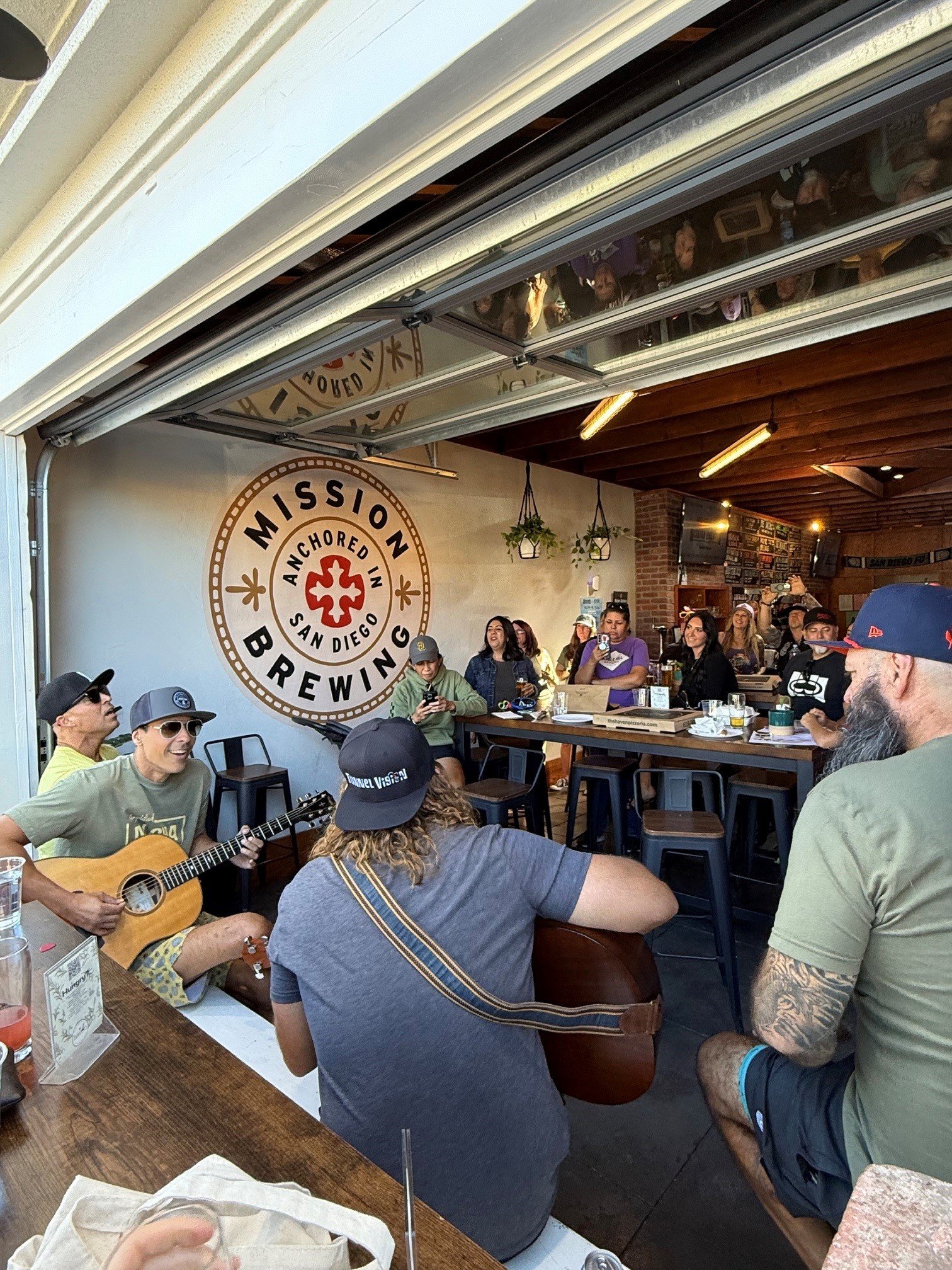 A group of people gathered indoors. Some are sitting, and others are standing or dancing. One person is playing a guitar. The wall features a large badge with the words 'Mission Brewing' and 'San Diego'. The ceiling is wooden with hanging plants. The