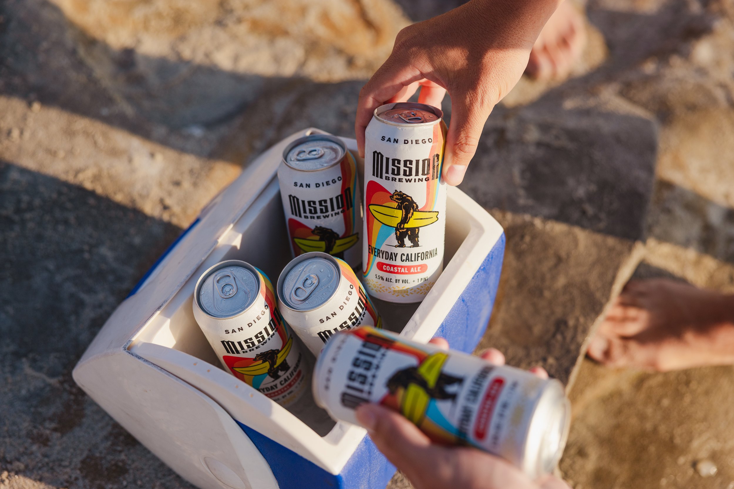 A person reaching for a can of Everyday California Mission Brewing beer into a white cooler in the sand. The can features a bear holding a surfboard with rainbow coloring, indicating a Coastal Ale with 5.5% alcohol, from San Diego, California.