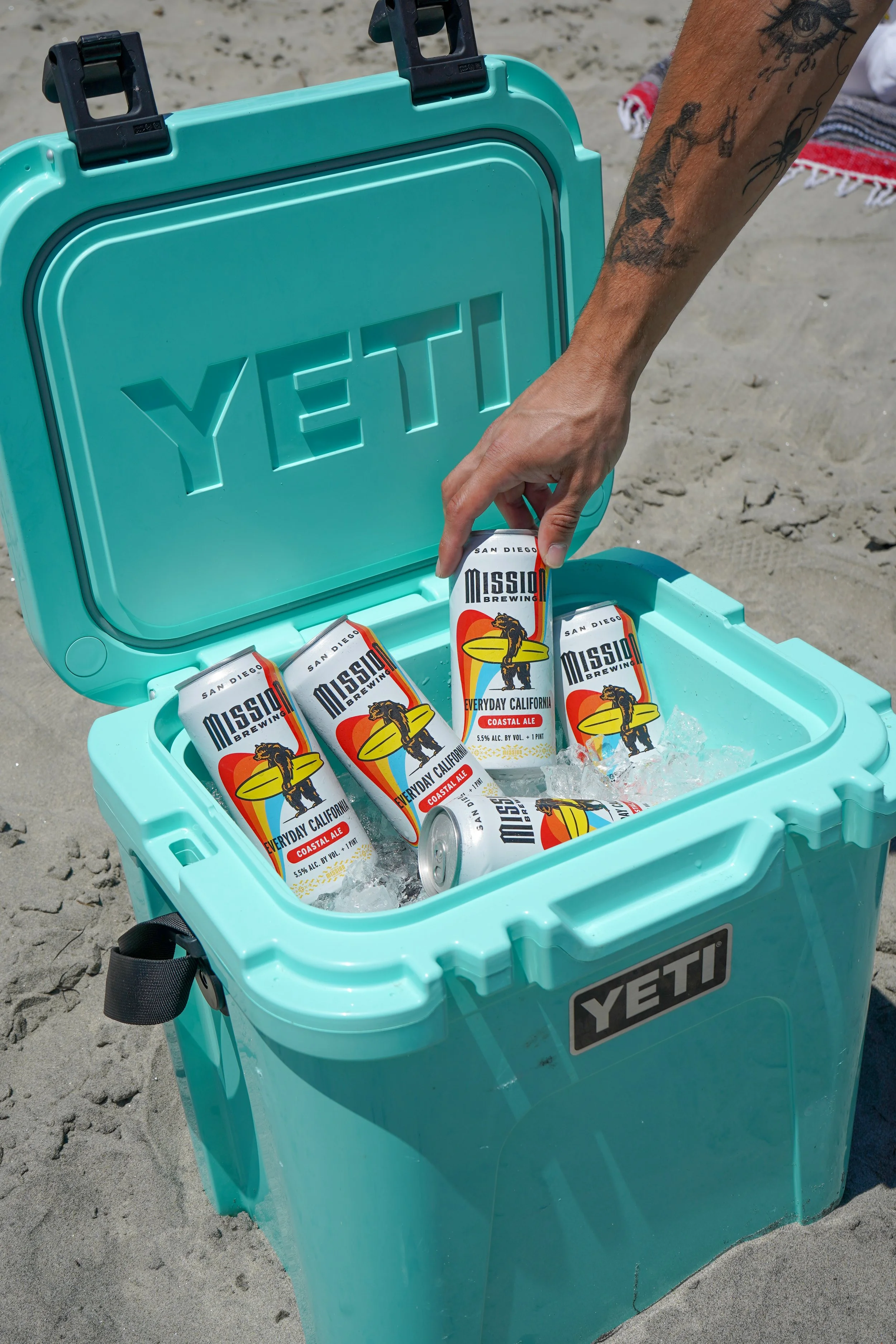 A turquoise YETI cooler filled with ice and cans of Mission Brewing Everyday California beer, with the lid open showing the cans inside, on a sandy beach.