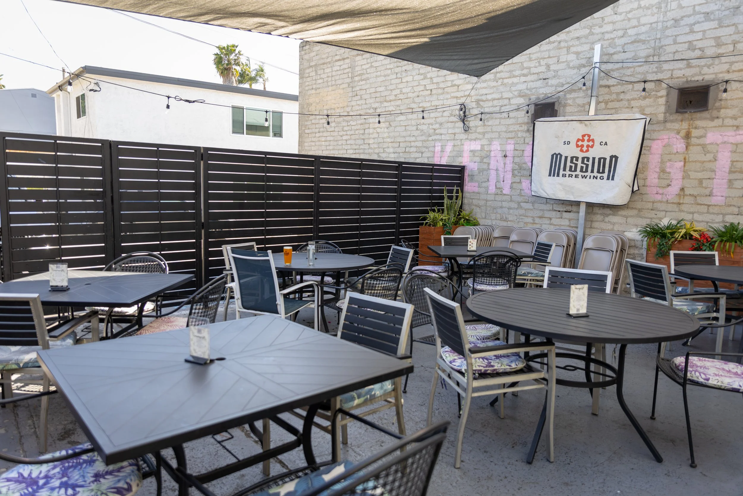 An outdoor back patio area at Mission Brewing Kensington with black tables and chairs, some with floral cushions, and potted plants. A brick wall with Kensington sign in pink graffiti with string lights hanging overhead.