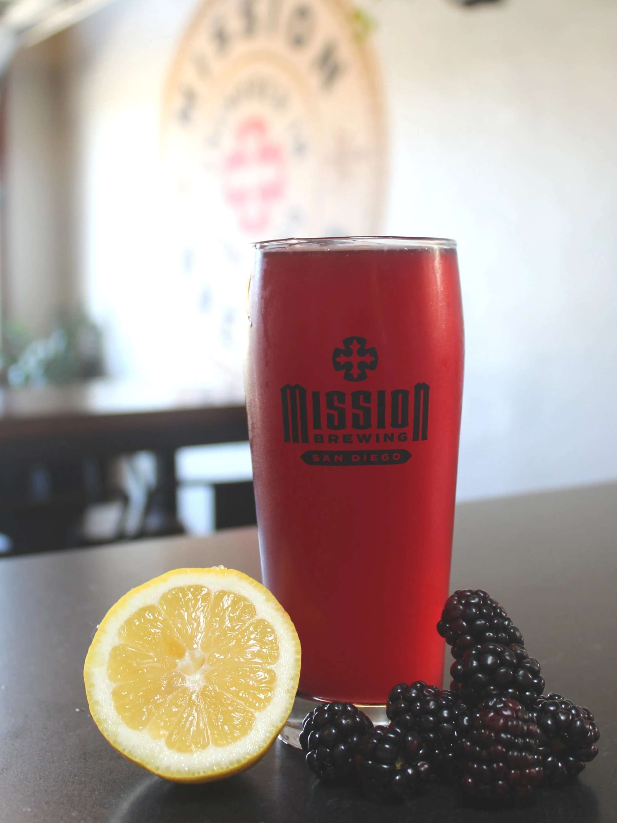 A Mission Brewing beer glass filled with red seltzer with blackberries and lemon slices on a dark surface indoors.