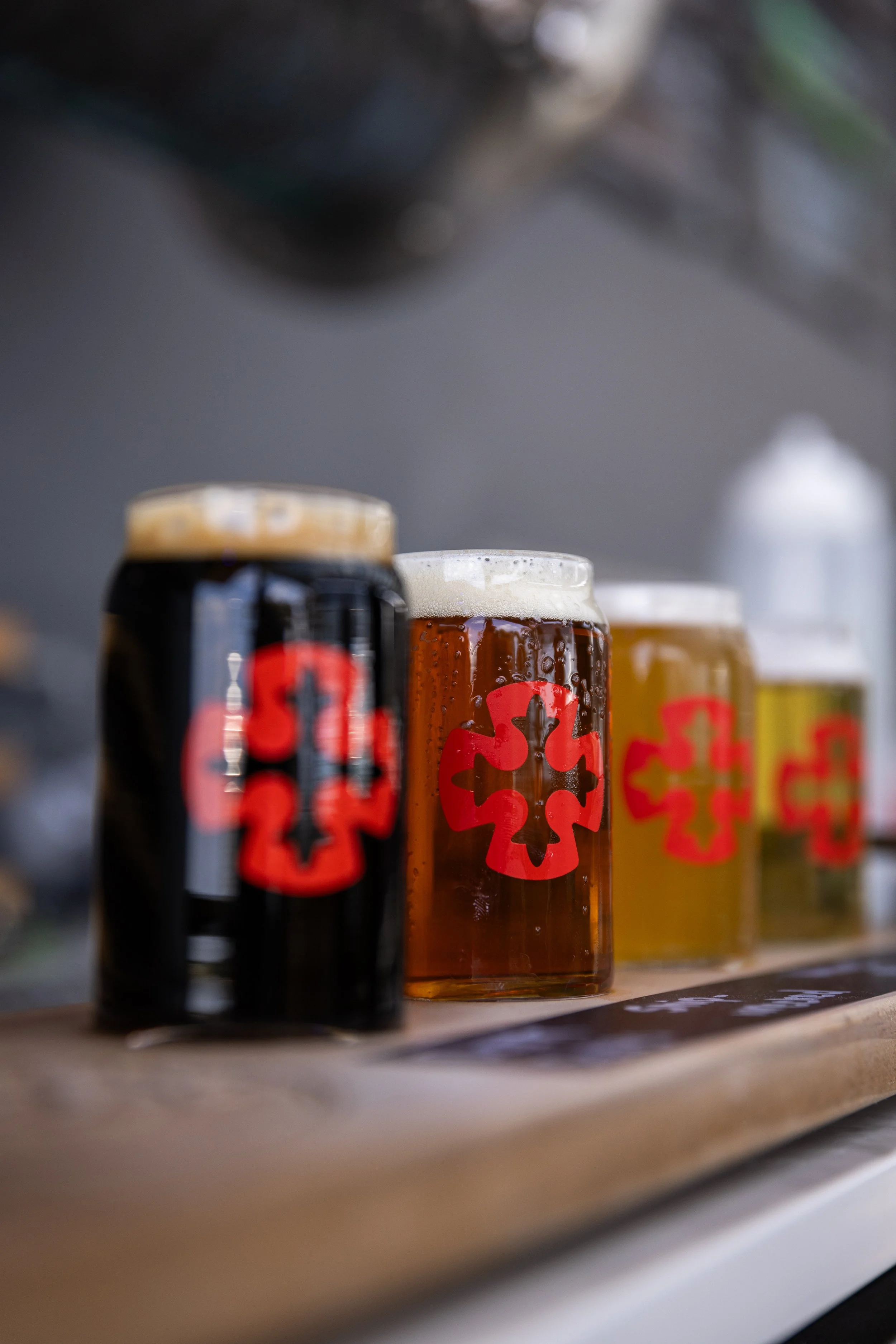 Four small glass cups with black, brown, and yellow colored draft beers on a wooden surface. Each jar has a red Mission Brewing logo.