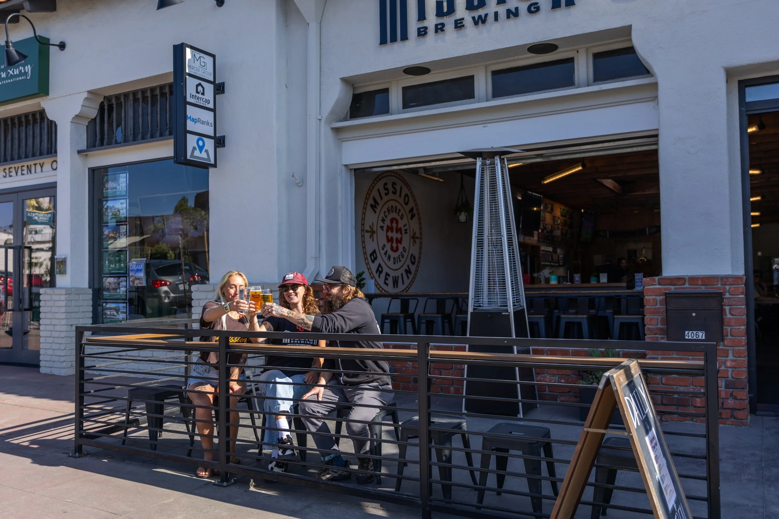 Three people sitting outdoors at a table raising glasses of beer in a toast in front of Mission Brewing Kensington. 